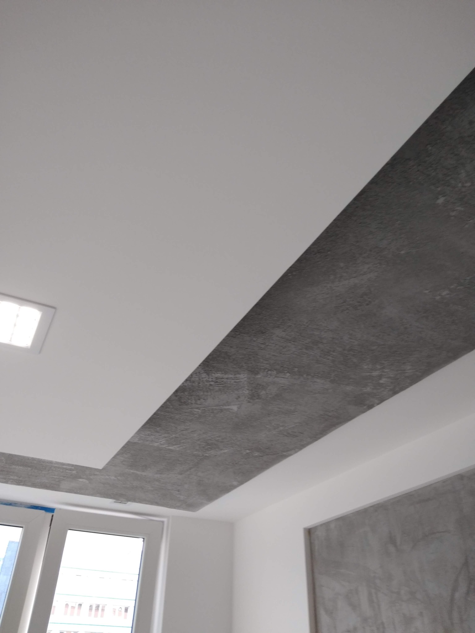 Fragment sufitu z dekoracyjnym pasem szpachlowanym o szarej, surowej fakturze, kontrastującym z gładką, białą powierzchnią sufitu i ścian, widoczne okno z niebieską taśmą zabezpieczającą.