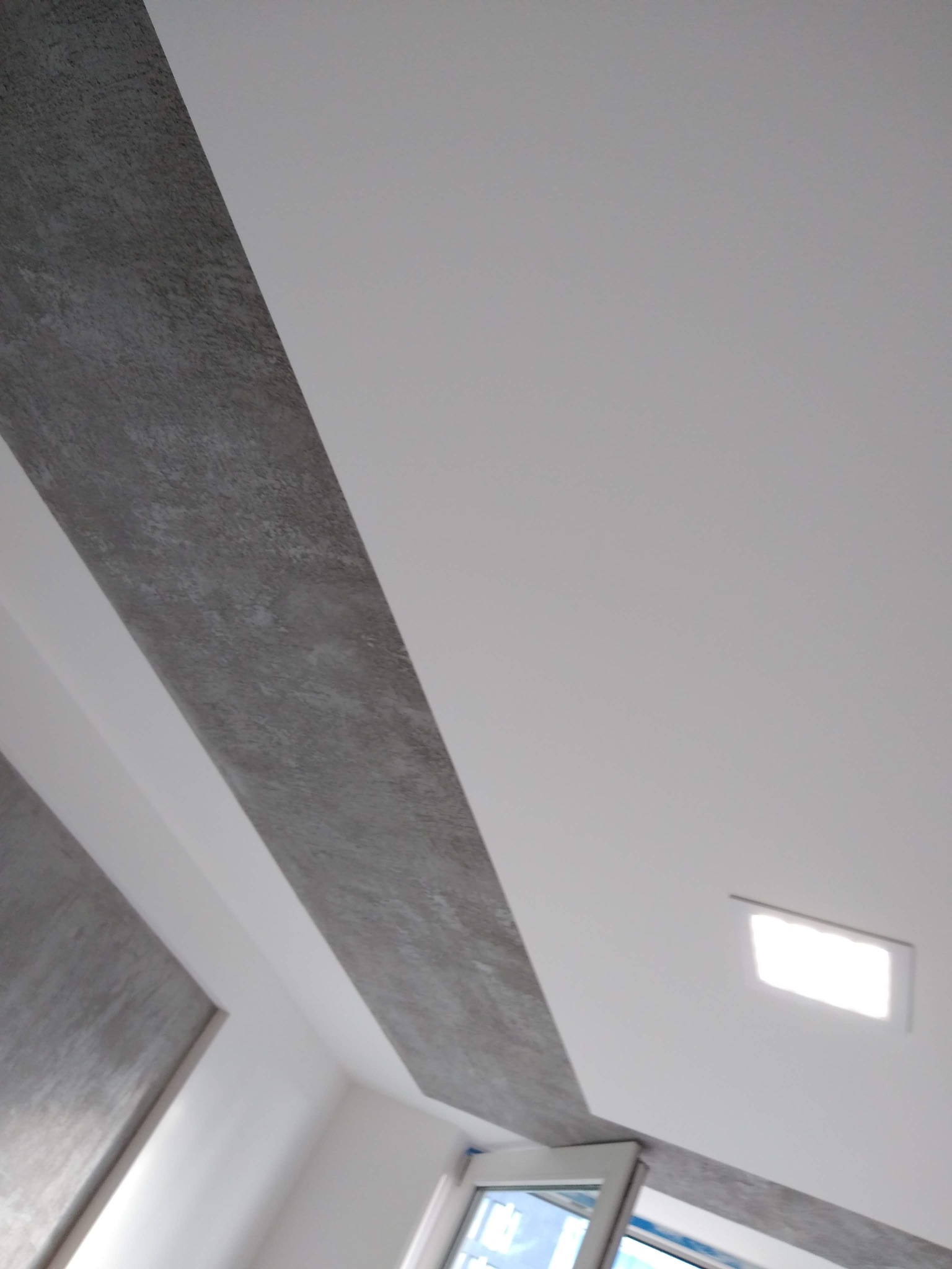 Wnętrze z wykończonym sufitem w biało-szare pasy, z wbudowanym oświetleniem LED i fragmentem okna w rogu.