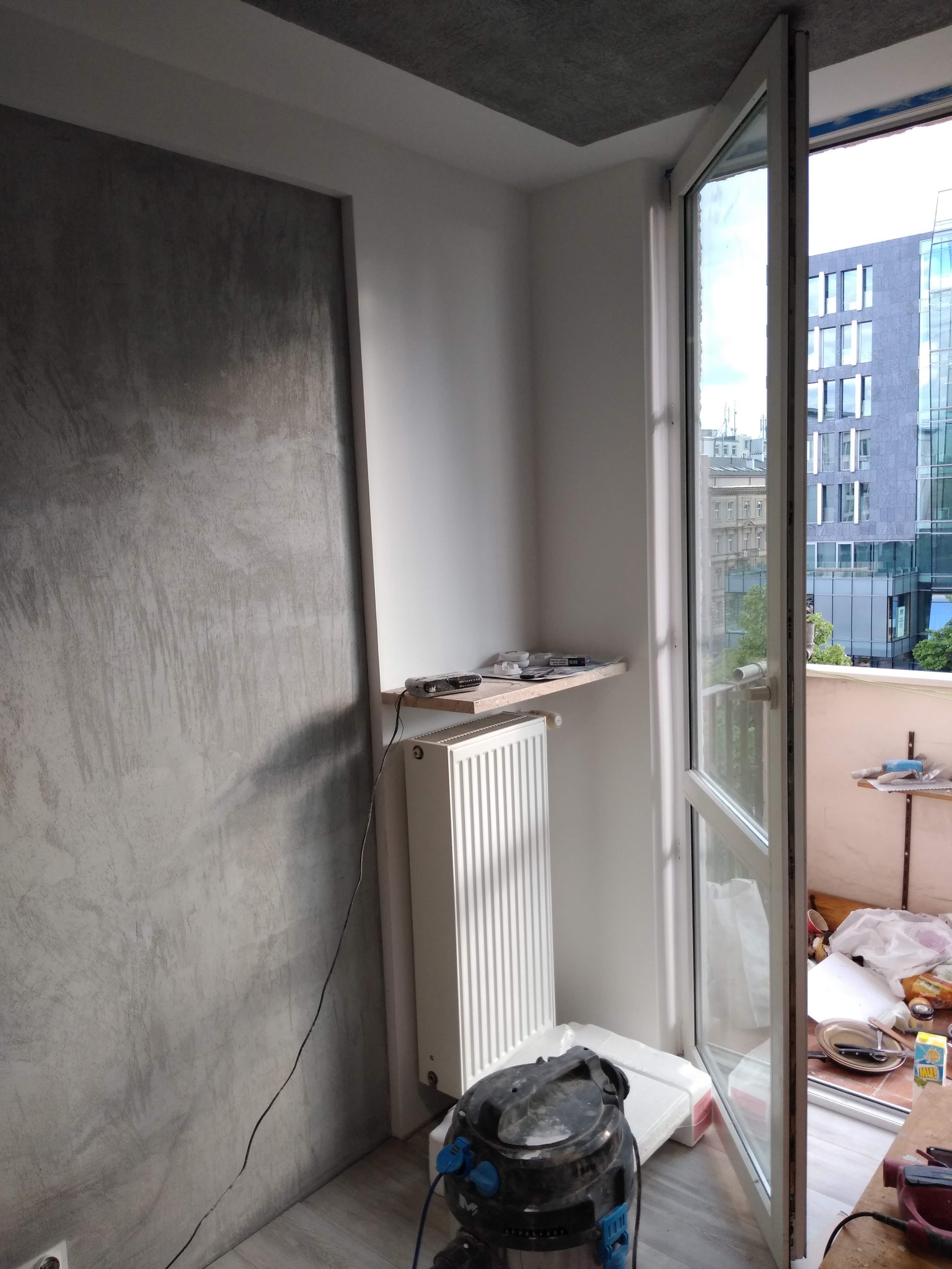 Ściana z efektem betonu architektonicznego w trakcie realizacji, widoczny grzejnik, półka z narzędziami i otwarte drzwi balkonowe z widokiem na miasto.