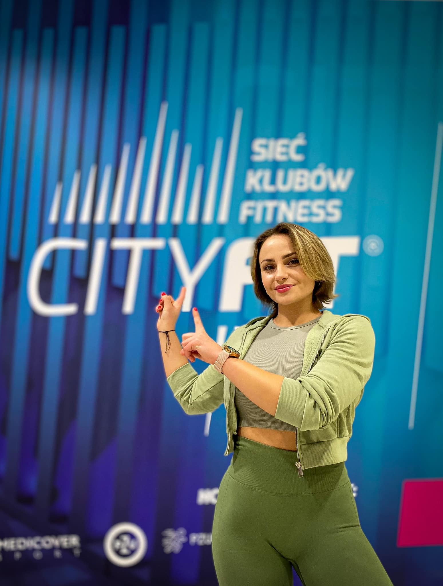 Kobieta w sportowym stroju wskazuje palcami na logo sieci klubów fitness Cityfit na niebieskim tle.