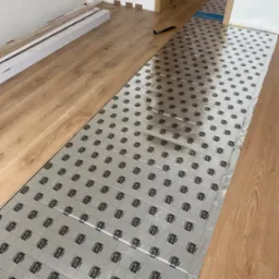 Proces układania paneli podłogowych: rozłożona folia izolacyjna z nadrukiem Cutting Lines, częściowo ułożone panele o jasnym odcieniu drewna, widoczne narzędzia i opakowania paneli w tle.