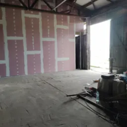 Wnętrze budynku w trakcie prac wykończeniowych: ściana z płyt kartonowo-gipsowych z widocznymi łączeniami szpachlowanymi, surowe drewniane elementy konstrukcyjne dachu, betonowa podłoga...