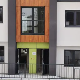 Nowoczesny budynek wielorodzinny w trakcie wykończenia, z elementami drewnopodobnymi i świeżo pomalowanym na jasnozielono wejściem do lokalu 48C/2.