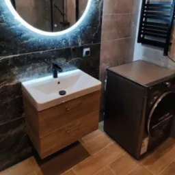 Nowoczesna łazienka z umywalką nablatową na drewnianej szafce, podświetlanym okrągłym lustrem i pralką LG w odcieniach szarości, wykończona płytkami imitującymi marmur i drewno.