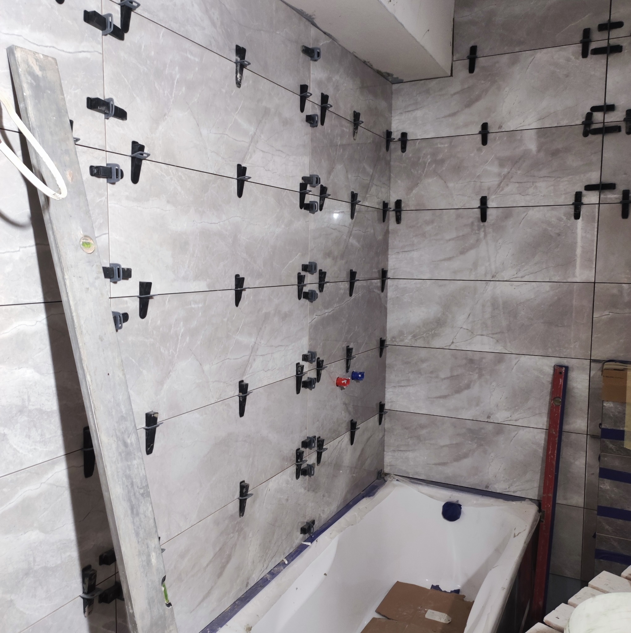 Montaż szarych płytek w łazience z użyciem klipsów poziomujących i krzyżyków dystansowych, widoczna wanna i poziomica.