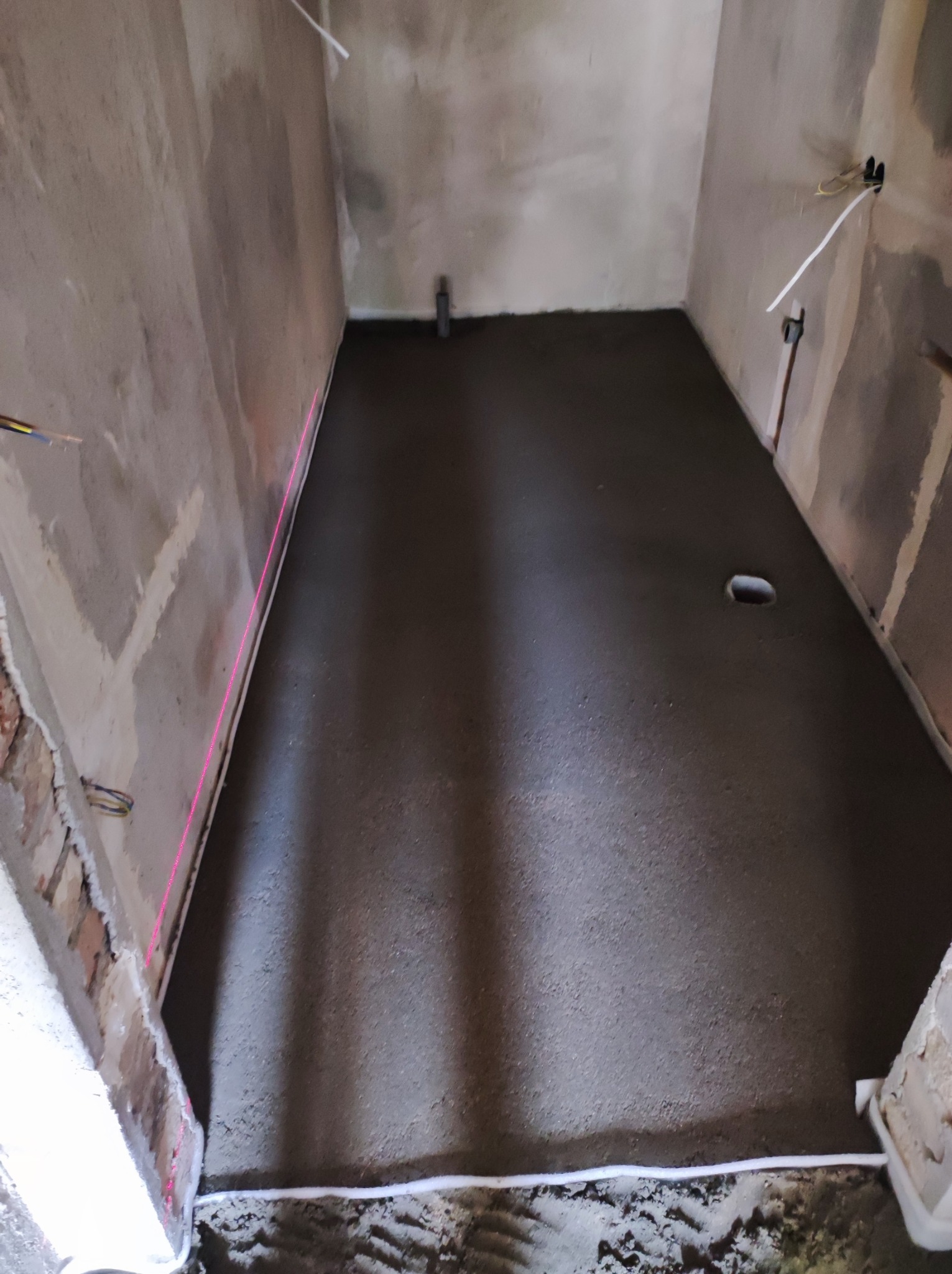 Wyrównana, szara wylewka podłogowa w pomieszczeniu z surowymi ścianami, widoczny laserowy poziom wyznaczający linię.