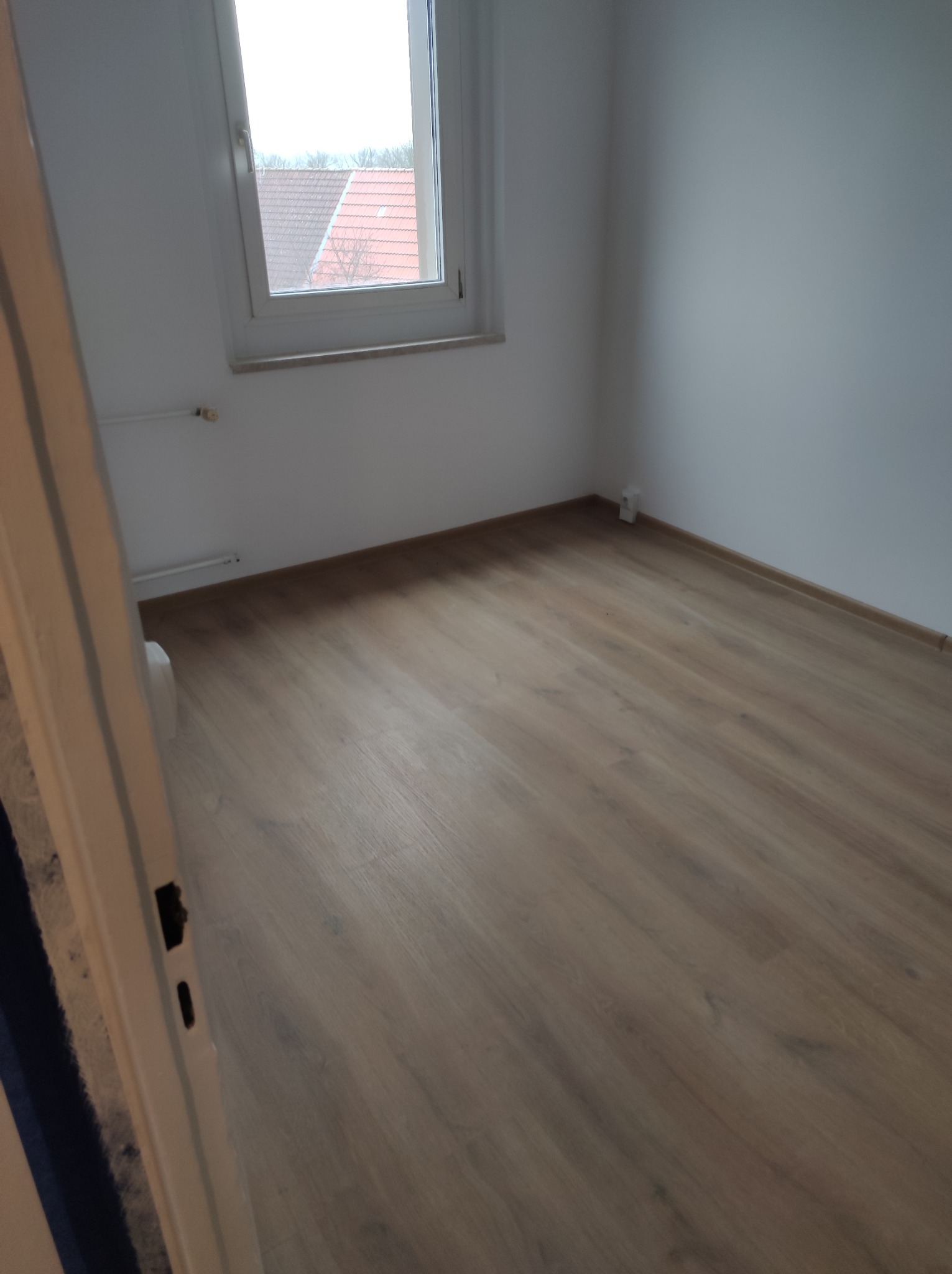 Puste, jasne pomieszczenie w Berlinku po remoncie, z panelami podłogowymi w odcieniach brązu i białymi ścianami. Widoczne okno z widokiem na dachy domów oraz fragment futryny drzwi po lewej stronie.