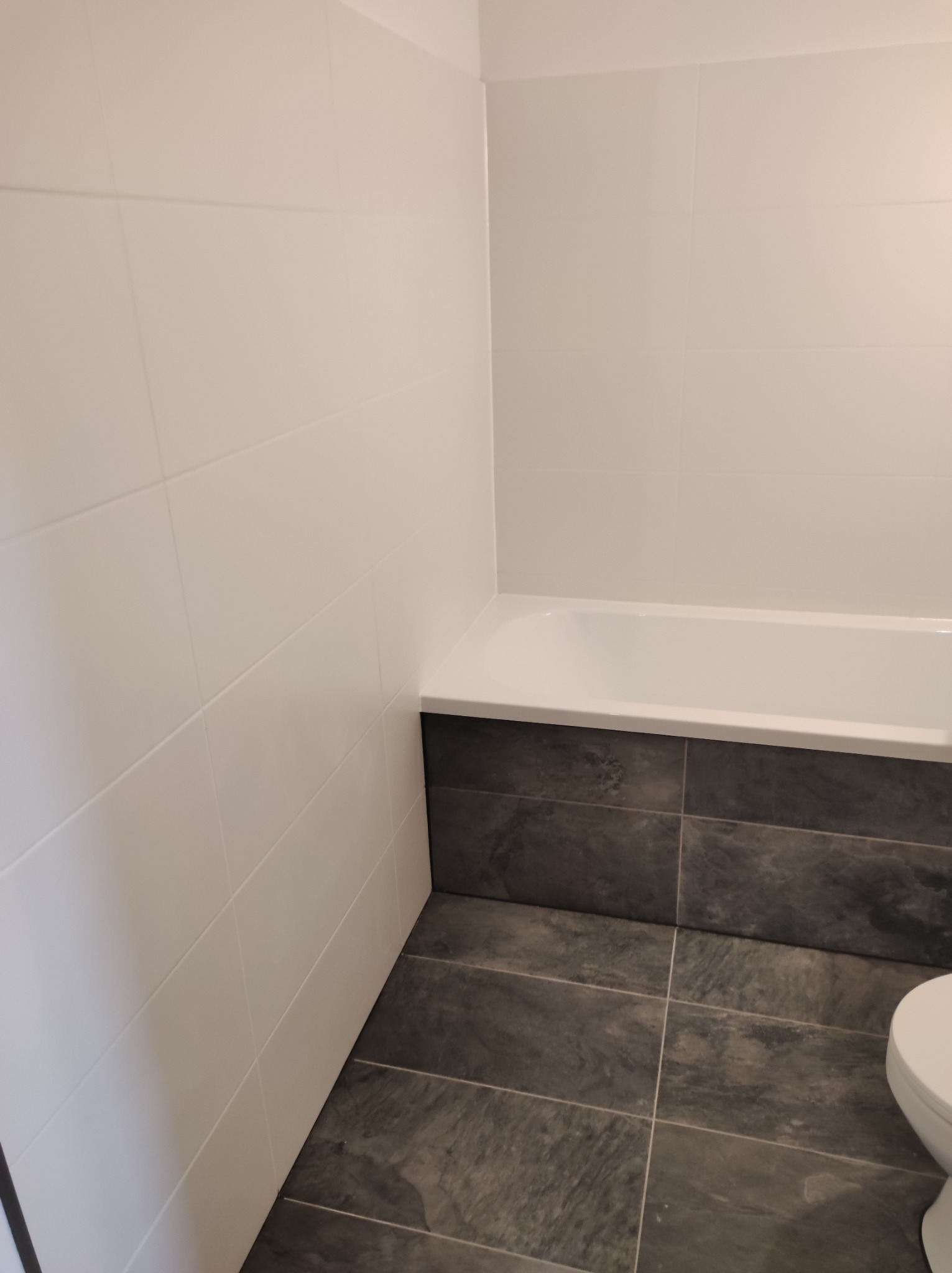 Wykończona łazienka z białymi płytkami na ścianach i ciemnoszarymi płytkami podłogowymi oraz obudową wanny, widoczny fragment toalety.