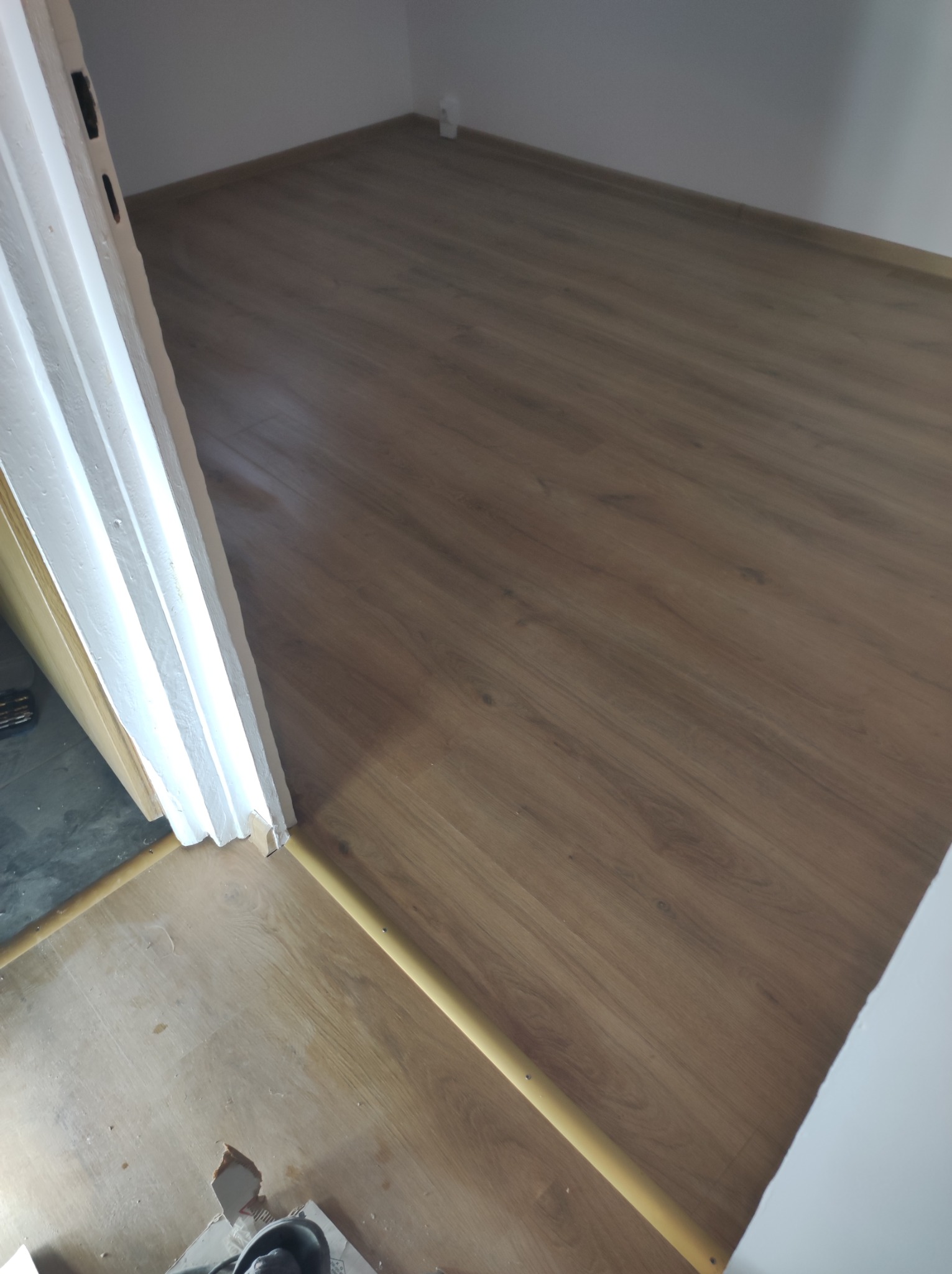 Ułożona podłoga z paneli w odcieniach brązu, widoczna listwa przypodłogowa i fragment ościeżnicy drzwiowej z widocznymi śladami prac wykończeniowych.