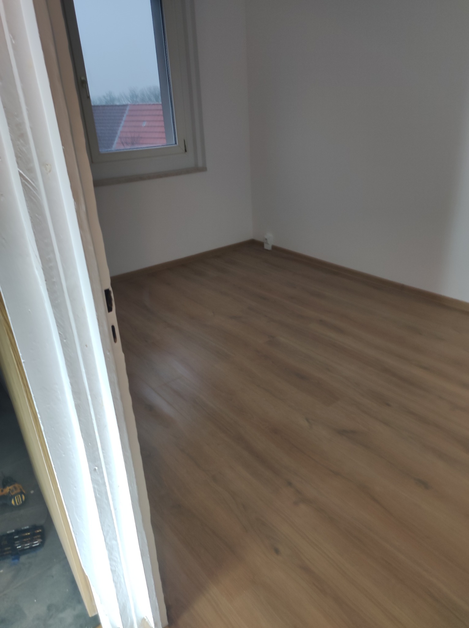 Pusty pokój po remoncie z nową drewnianą podłogą, świeżo pomalowanymi białymi ścianami i oknem z widokiem na szare niebo i dachy sąsiednich budynków. Częściowo widoczne drzwi po lewej stronie.