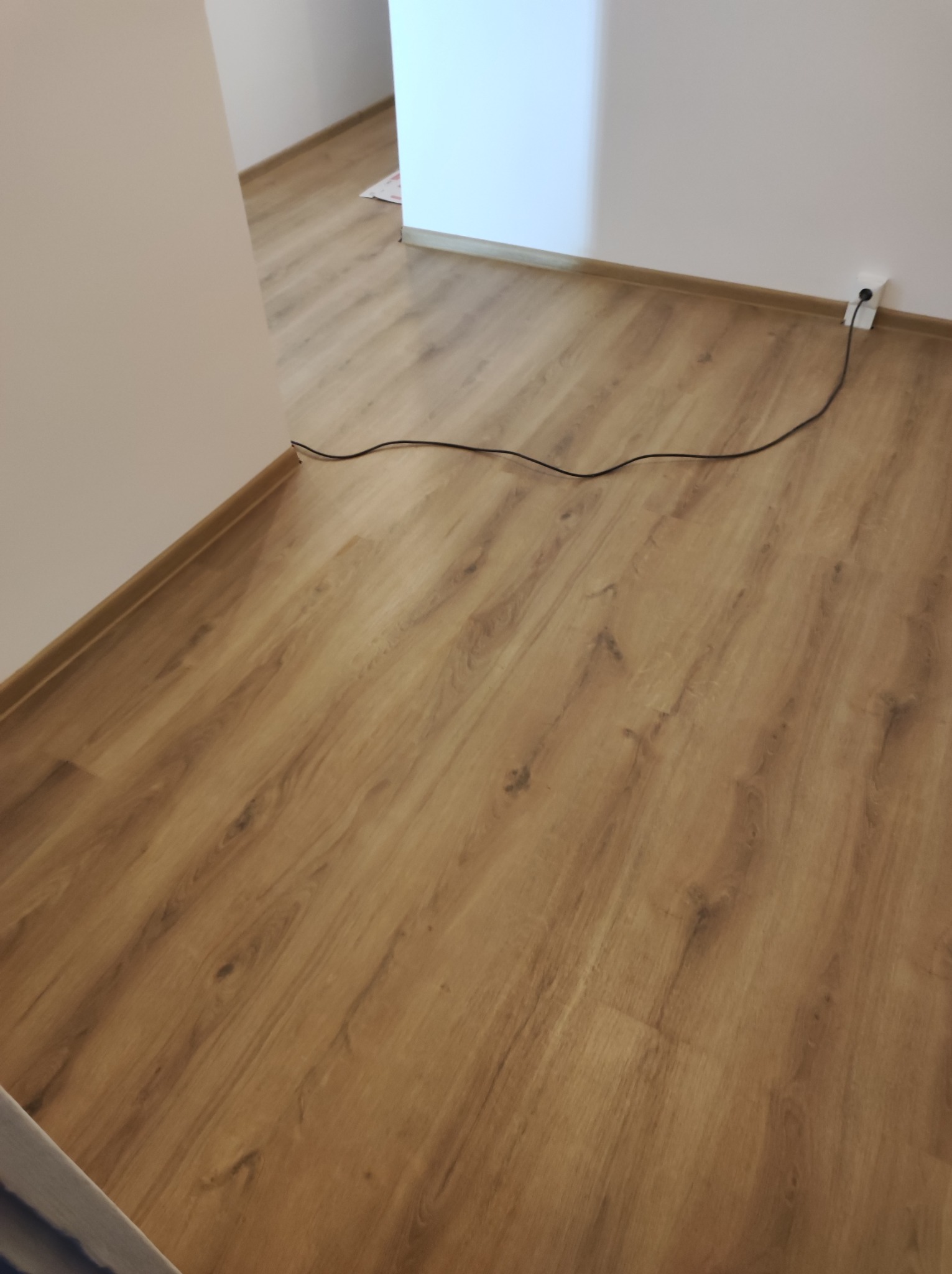 Nowa podłoga z paneli w odcieniach brązu, widoczna listwa przypodłogowa i kabel elektryczny przy ścianie.