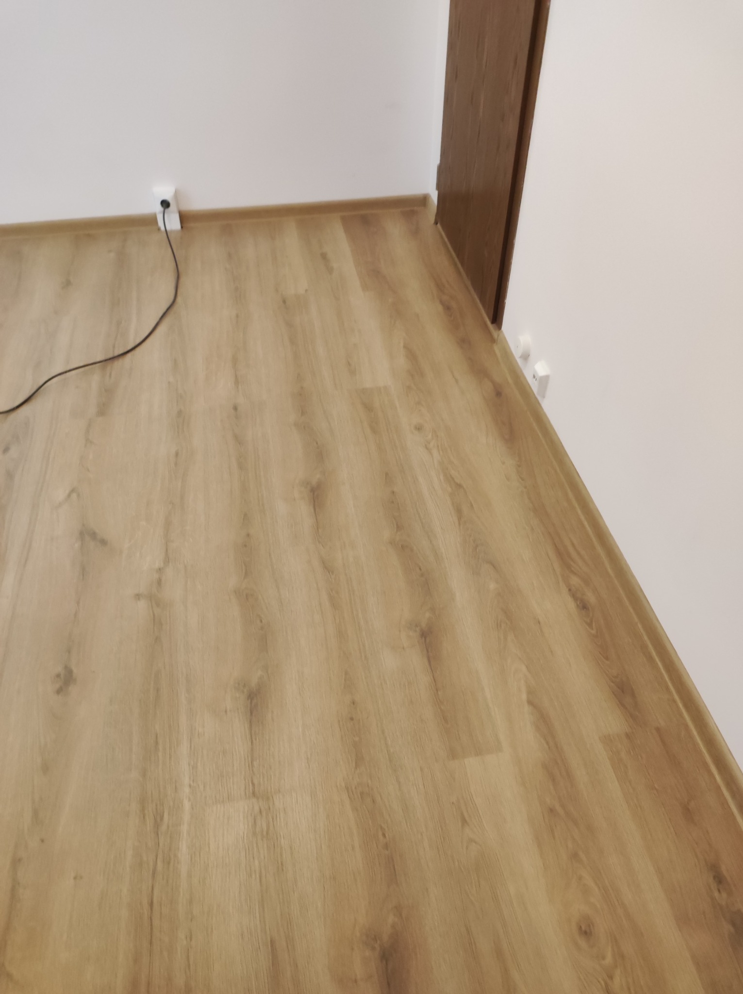 Jasna, drewnopodobna podłoga, widoczne listwy przypodłogowe przy białej ścianie, gniazdka elektryczne i fragment kabla w rogu pomieszczenia.