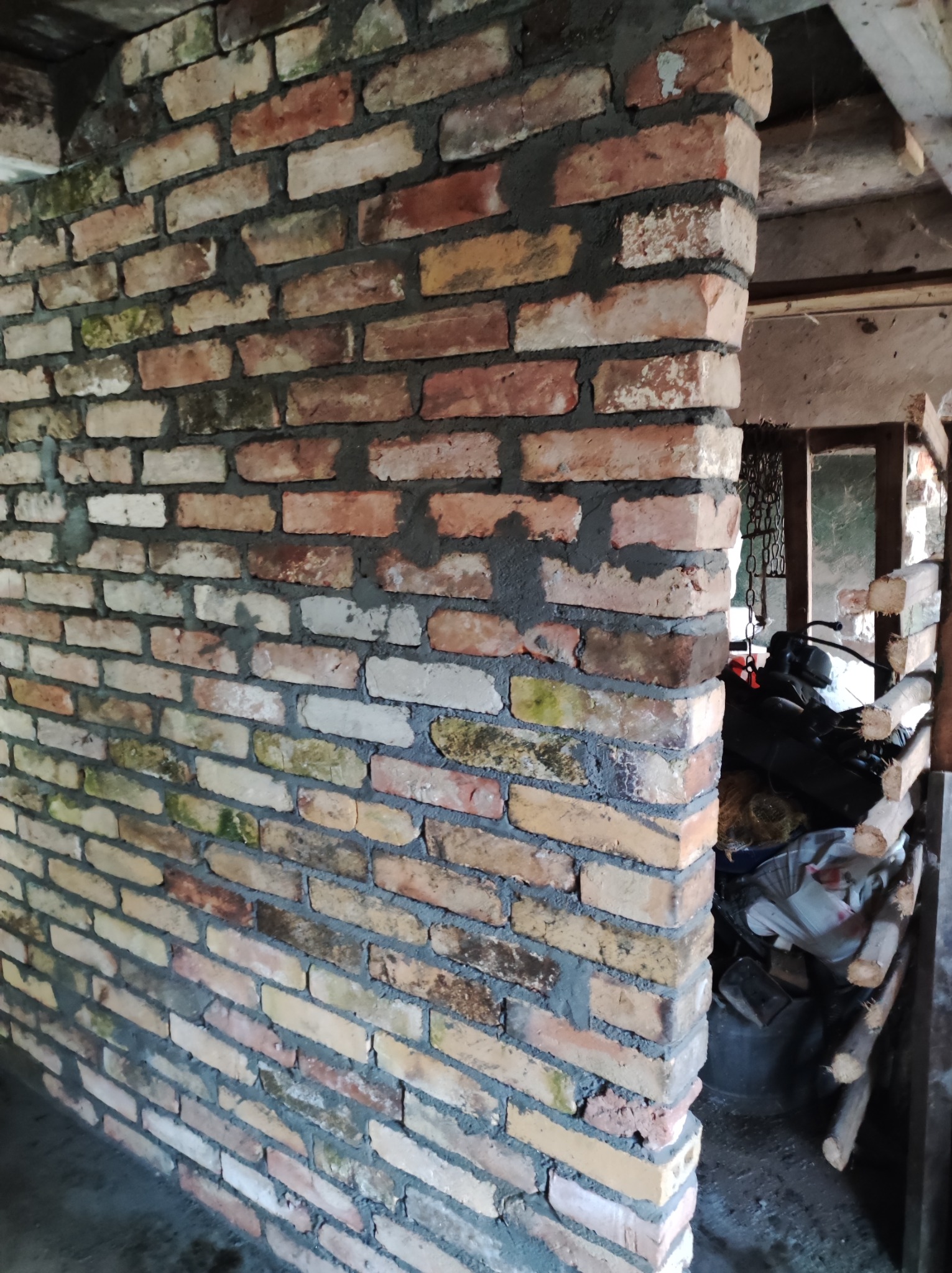 Murowana ściana z cegły wewnątrz budynku, widoczne ślady wilgoci i wykwitów na niektórych cegłach, surowe wykończenie z widocznym zapleczem.