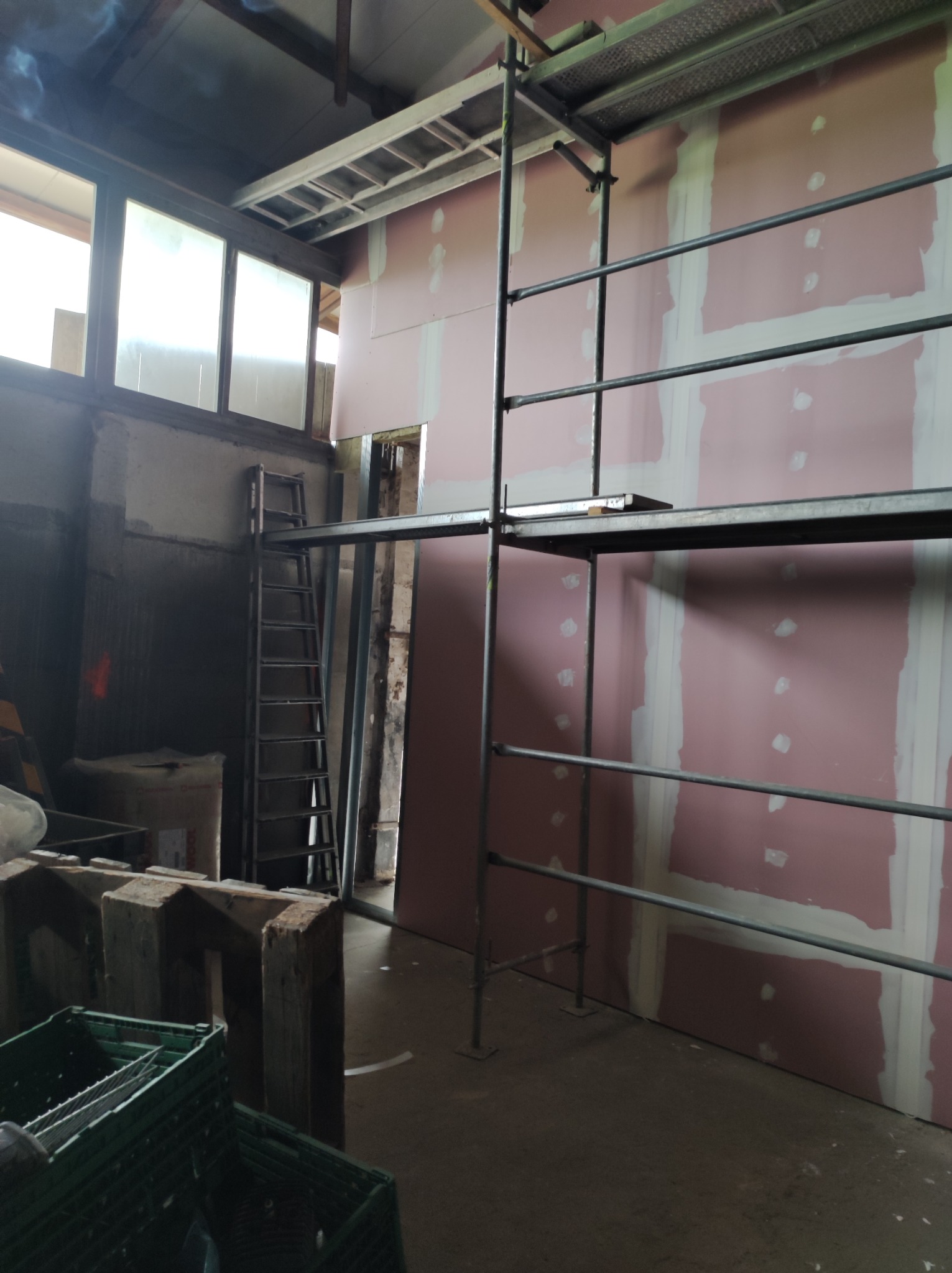 Montaż płyt karton gips na ścianie w surowym wnętrzu, widoczne rusztowanie i drabina, szpachlowane łączenia płyt.