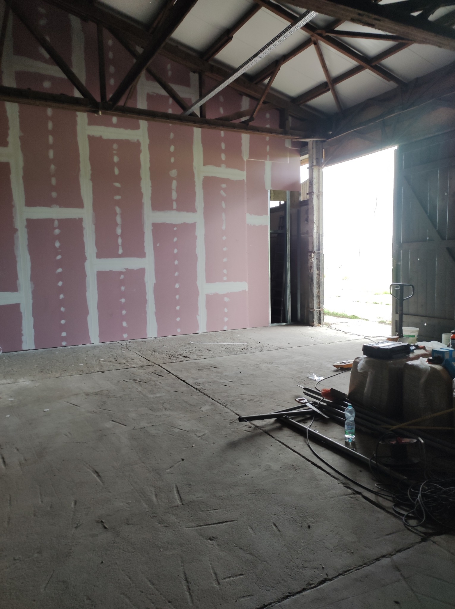 Wnętrze budynku w trakcie prac wykończeniowych: ściana z płyt kartonowo-gipsowych z widocznymi łączeniami szpachlowanymi, surowe drewniane elementy konstrukcyjne dachu, betonowa podłoga...