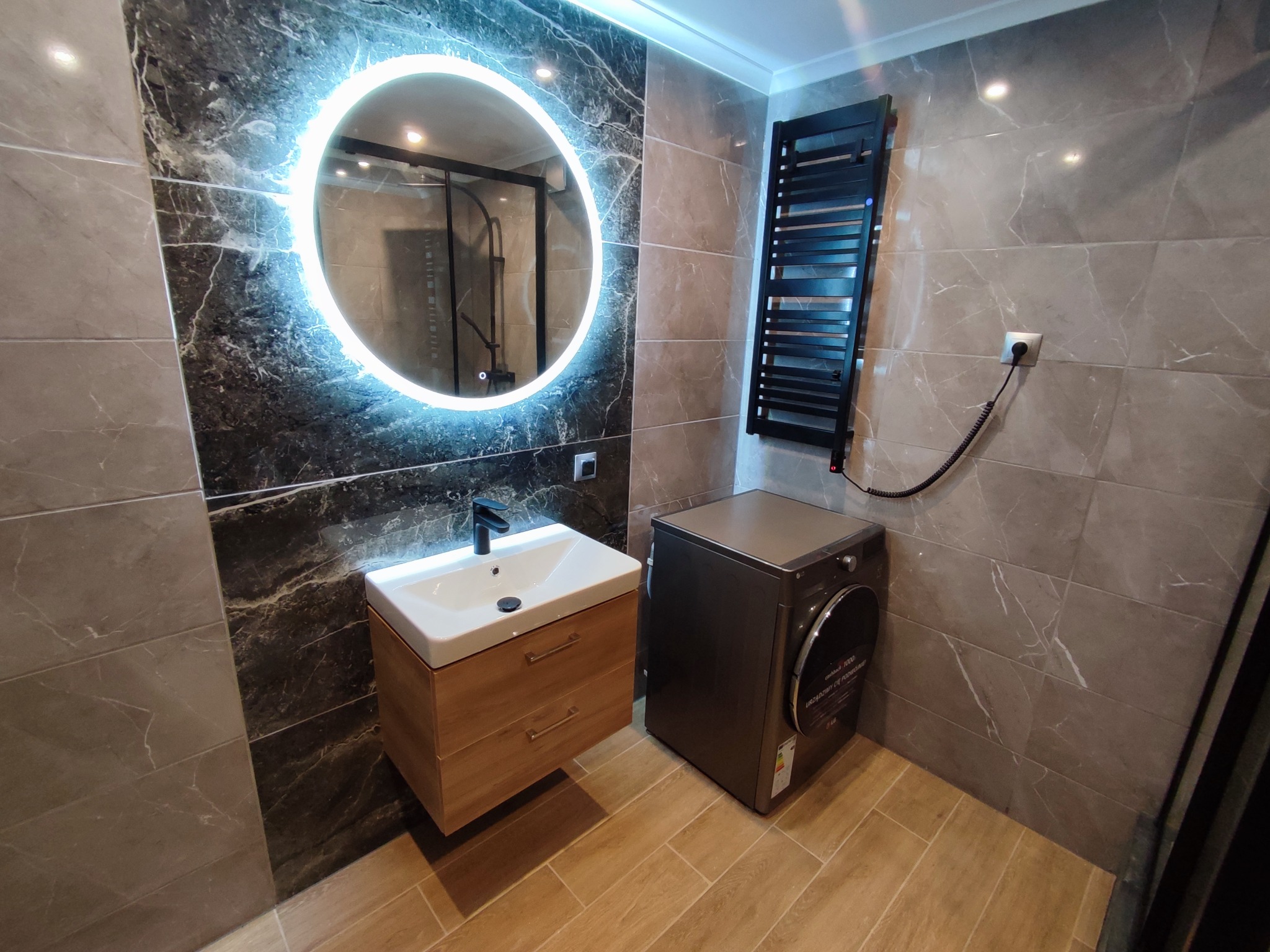 Nowoczesna łazienka z marmurowymi płytkami, okrągłym lustrem z podświetleniem LED, drewnianą szafką pod umywalką, grzejnikiem drabinkowym i pralko-suszarką LG.