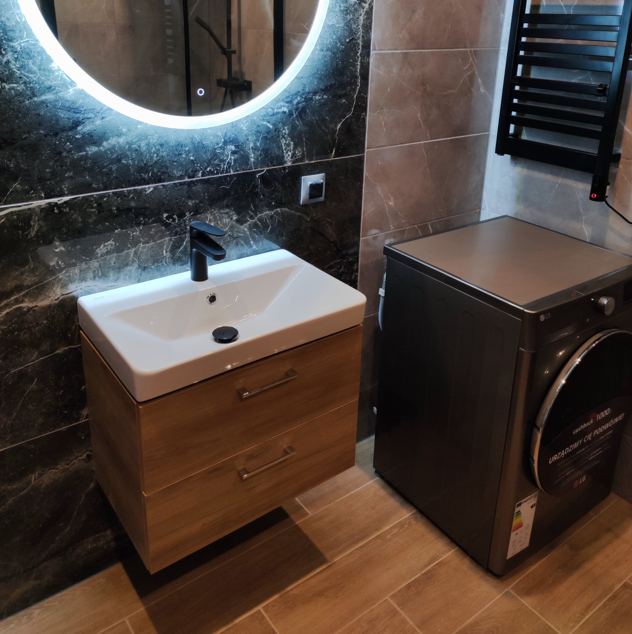 Nowoczesna łazienka z umywalką nablatową na drewnianej szafce, podświetlanym okrągłym lustrem i pralką LG w odcieniach szarości, wykończona płytkami imitującymi marmur i drewno.