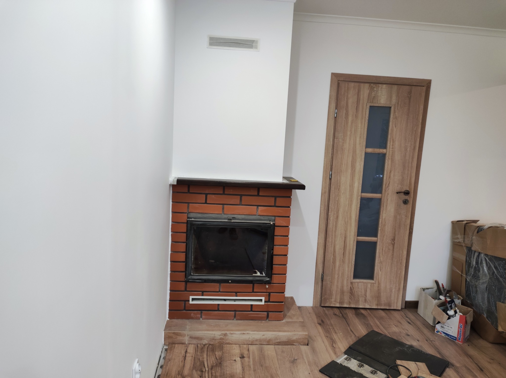Ceglany kominek z czarnym wkładem, umieszczony przy białej ścianie z kratką wentylacyjną, na drewnianej podłodze z widocznymi elementami wykończeniowymi i drzwiami w tle.