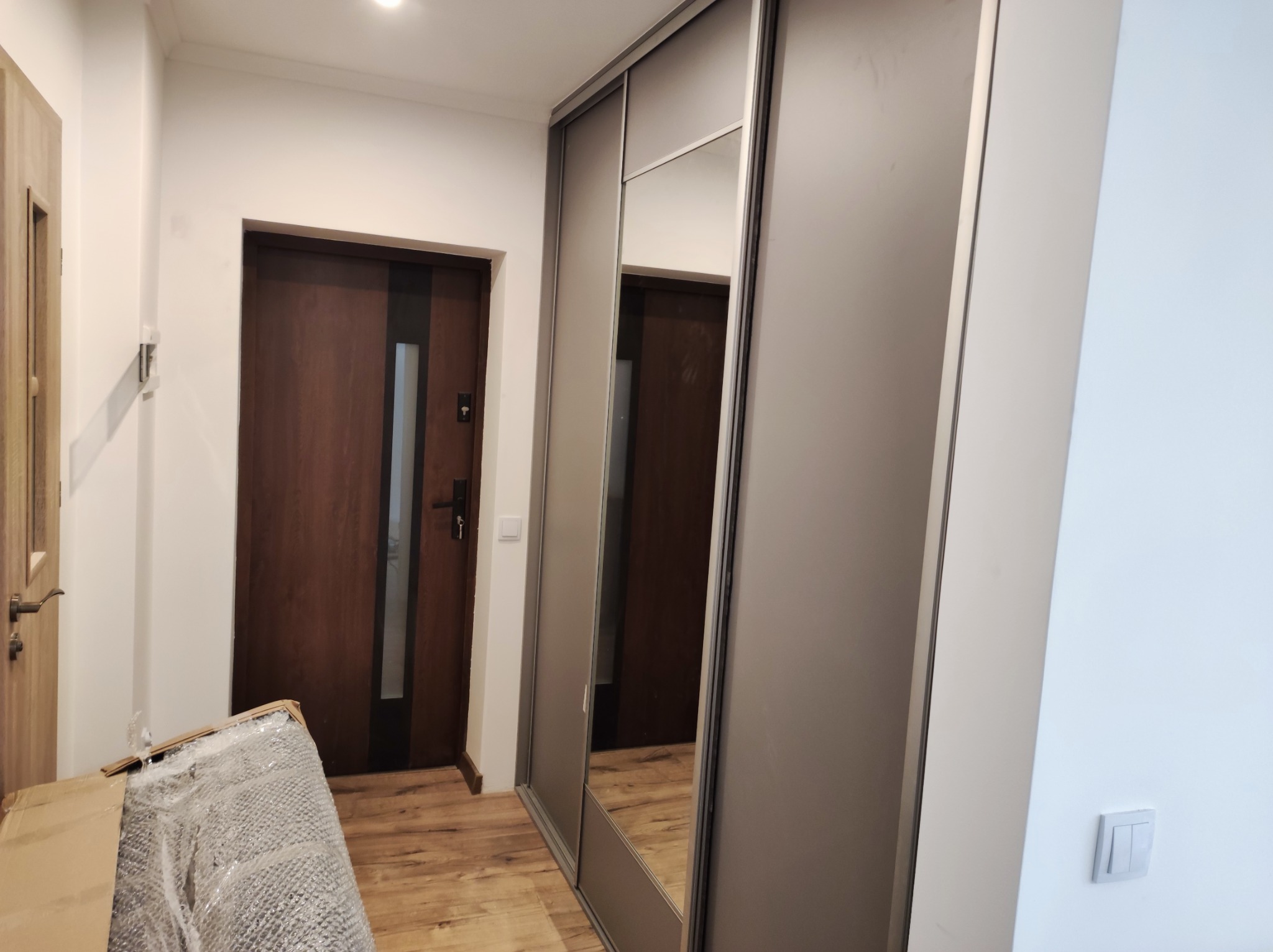 Korytarz po remoncie z dużą szafą przesuwną z lustrem, widoczne drzwi wejściowe i drzwi wewnętrzne, podłoga drewniana, fragment kartonu zabezpieczającego na podłodze.
