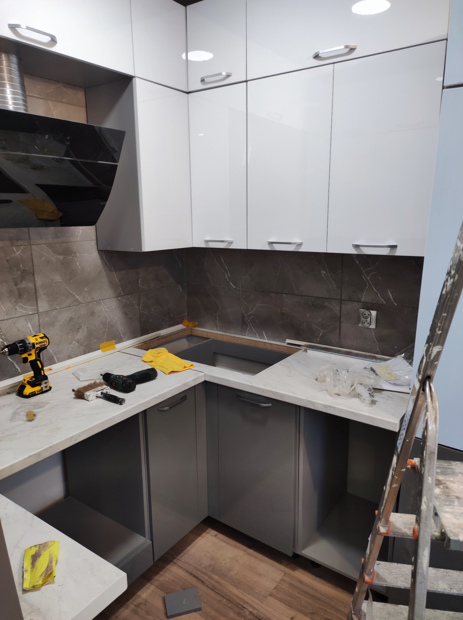 Szaro-biała kuchnia w trakcie montażu: widoczne szafki górne i dolne, płytki imitujące marmur na ścianie, drabina oparta o ścianę, narzędzia (wiertarka, śrubokręt) i akcesoria na blacie przed...