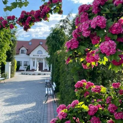 Ujęcie z zewnątrz budynku z nazwą 'Biały Dworek', otoczonego kwitnącymi krzewami i zadbanym ogrodem z alejką i ławeczkami.