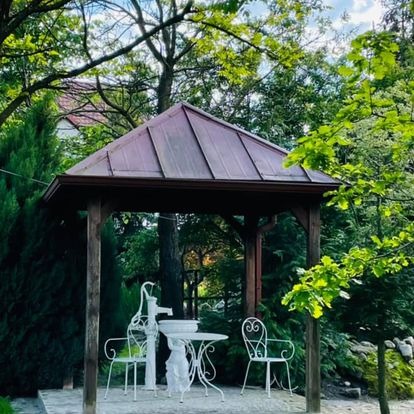 Altana ogrodowa z drewnianą konstrukcją i rdzawym dachem, wyposażona w biały, ażurowy stolik i krzesła, z ozdobną pompą wodną po lewej stronie, otoczona zielenią drzew i krzewów.