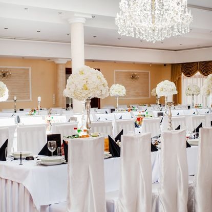 Sala bankietowa z ustawionymi stołami na uroczystość: białe obrusy, krzesła z pokrowcami, czarne serwetki, szklanki, talerze i wysokie kompozycje kwiatowe w srebrnych wazonach, oświetlona...