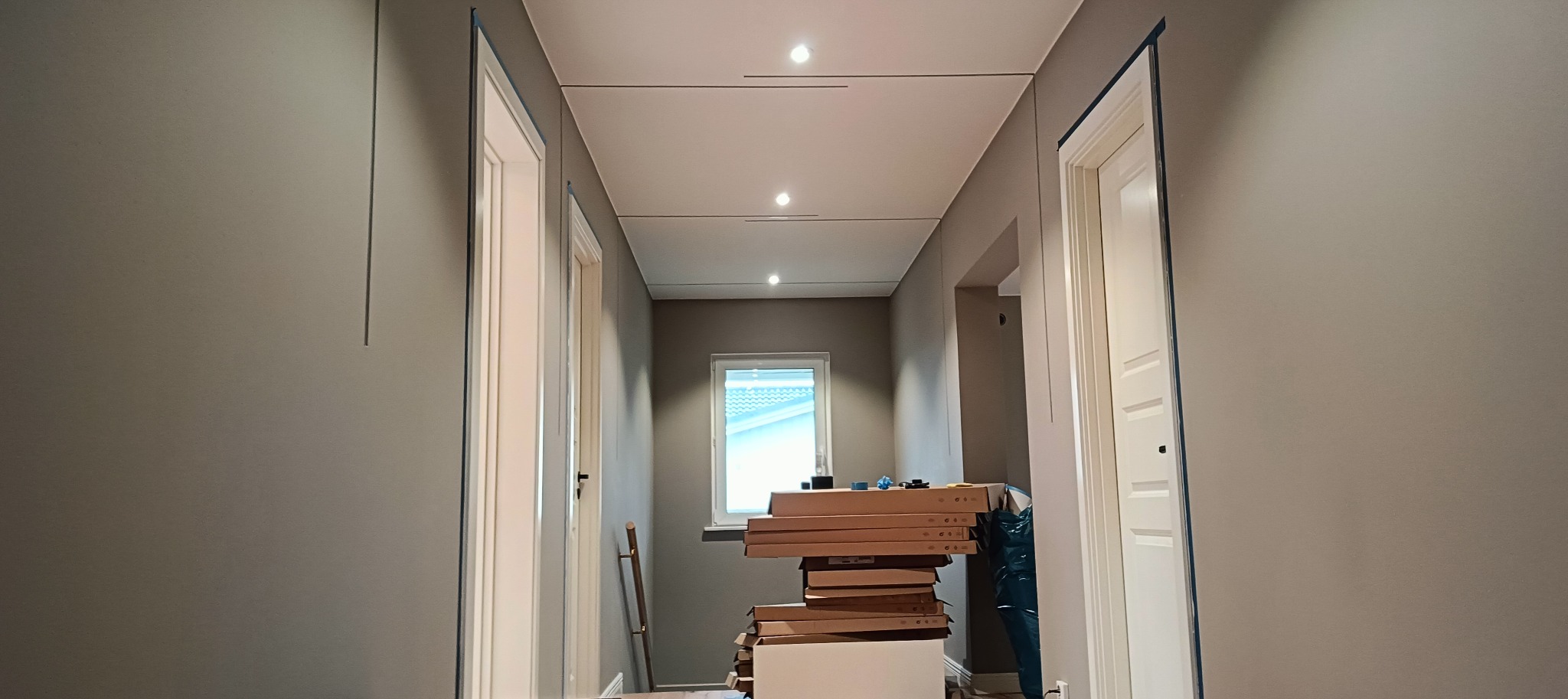 Korytarz w trakcie przygotowań do malowania, ściany w szarym odcieniu z niebieską taśmą malarską przy białych futrynach drzwi, na podłodze stos kartonów, sufit z oświetleniem punktowym.