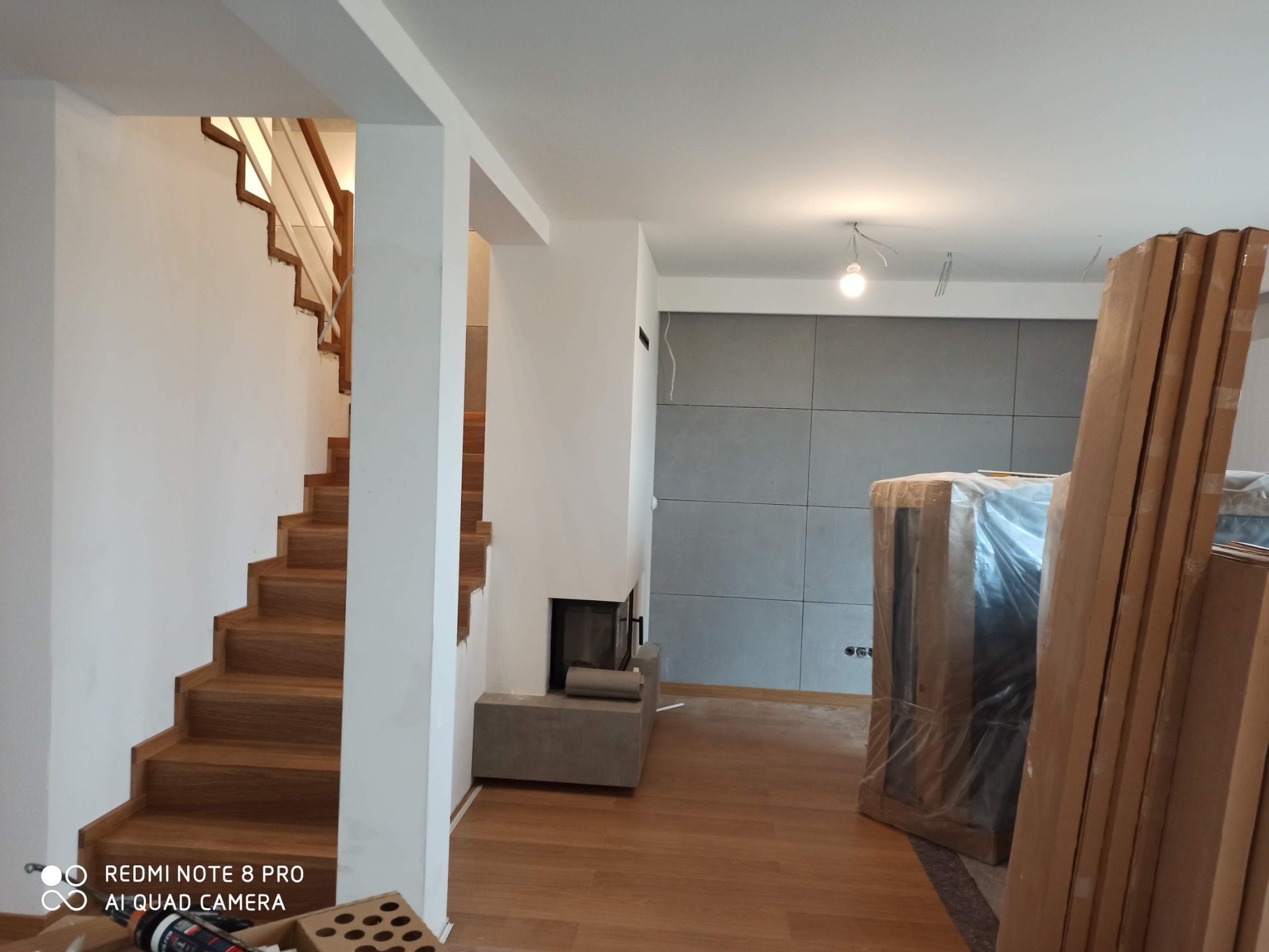 Wnętrze w trakcie prac wykończeniowych: drewniane schody, ściana z szarych paneli, obudowa kominka z betonu architektonicznego oraz elementy wyposażenia zabezpieczone folią i kartonem.