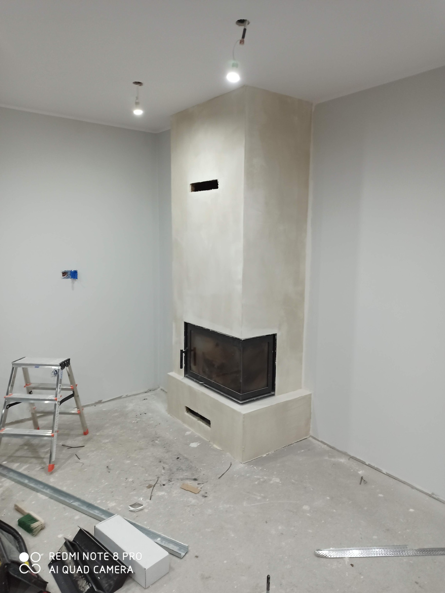 Wnętrze w trakcie prac: otynkowany kominek z czarnym wkładem kominkowym, drabina aluminiowa, profile metalowe i narzędzia na zabezpieczonej podłodze, surowe żarówki pod sufitem.