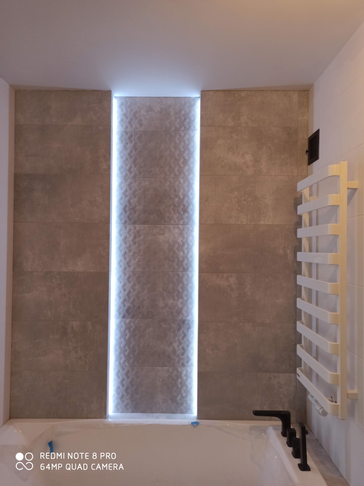 Wykończona łazienka z podświetlaną wnęką z dekoracyjnymi płytkami, grzejnik drabinkowy i czarna armatura.