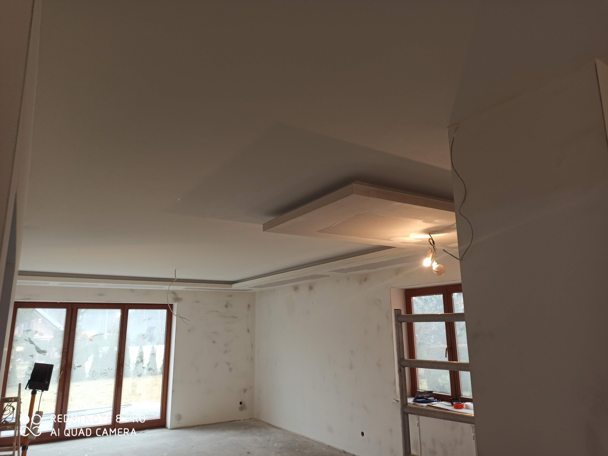 Pokój w trakcie prac przygotowawczych do malowania: surowe ściany z widocznymi śladami szpachlowania, podwieszany sufit z płyt kartonowo-gipsowych, widoczne kable oświetleniowe, drabina aluminiowa...