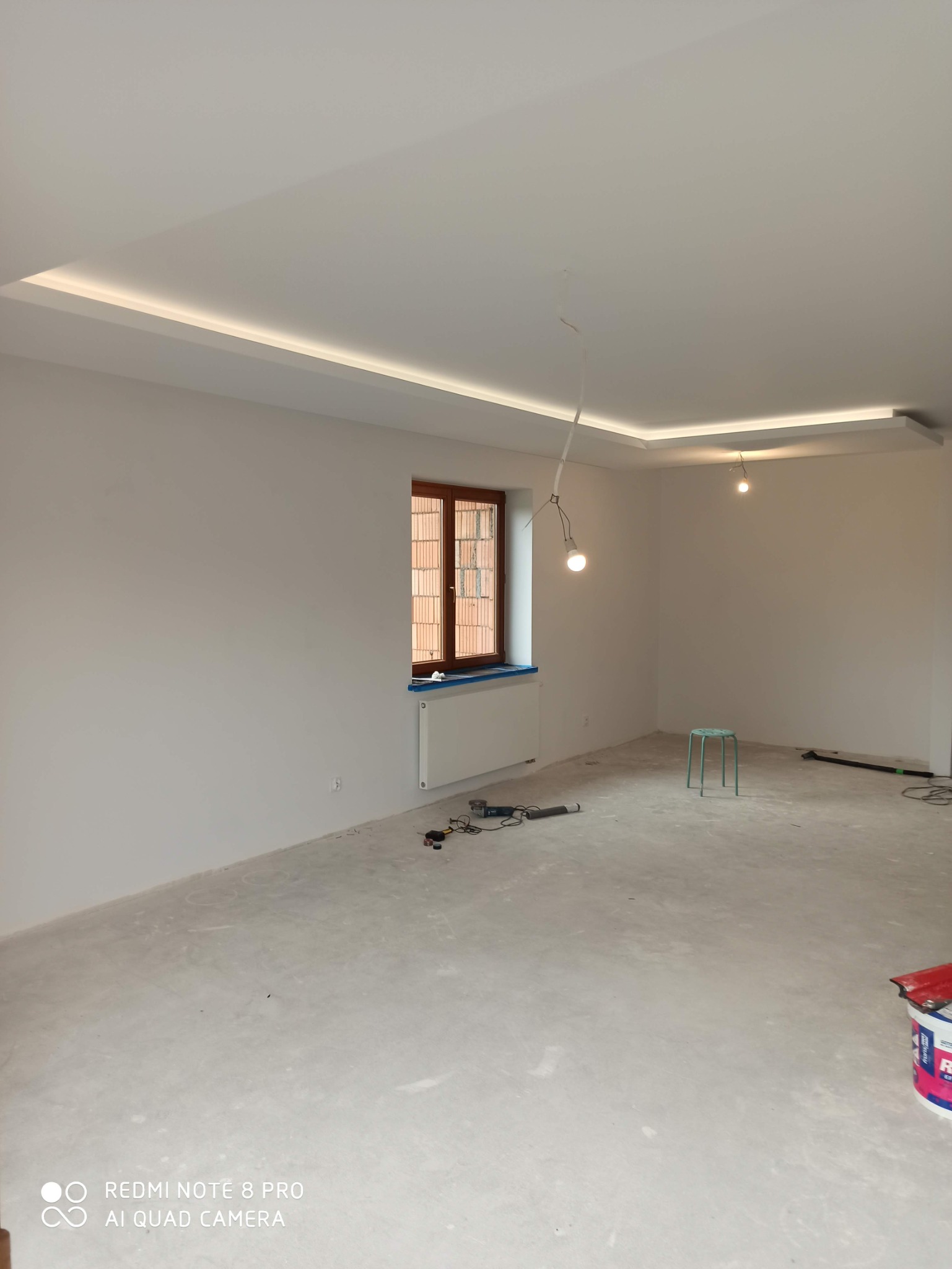 Pokój w trakcie prac wykończeniowych, z widoczną surową podłogą, świeżo pomalowanymi ścianami, oknem z drewnianą ramą, grzejnikiem, narzędziami na podłodze i minimalistycznym oświetleniem sufitowym.