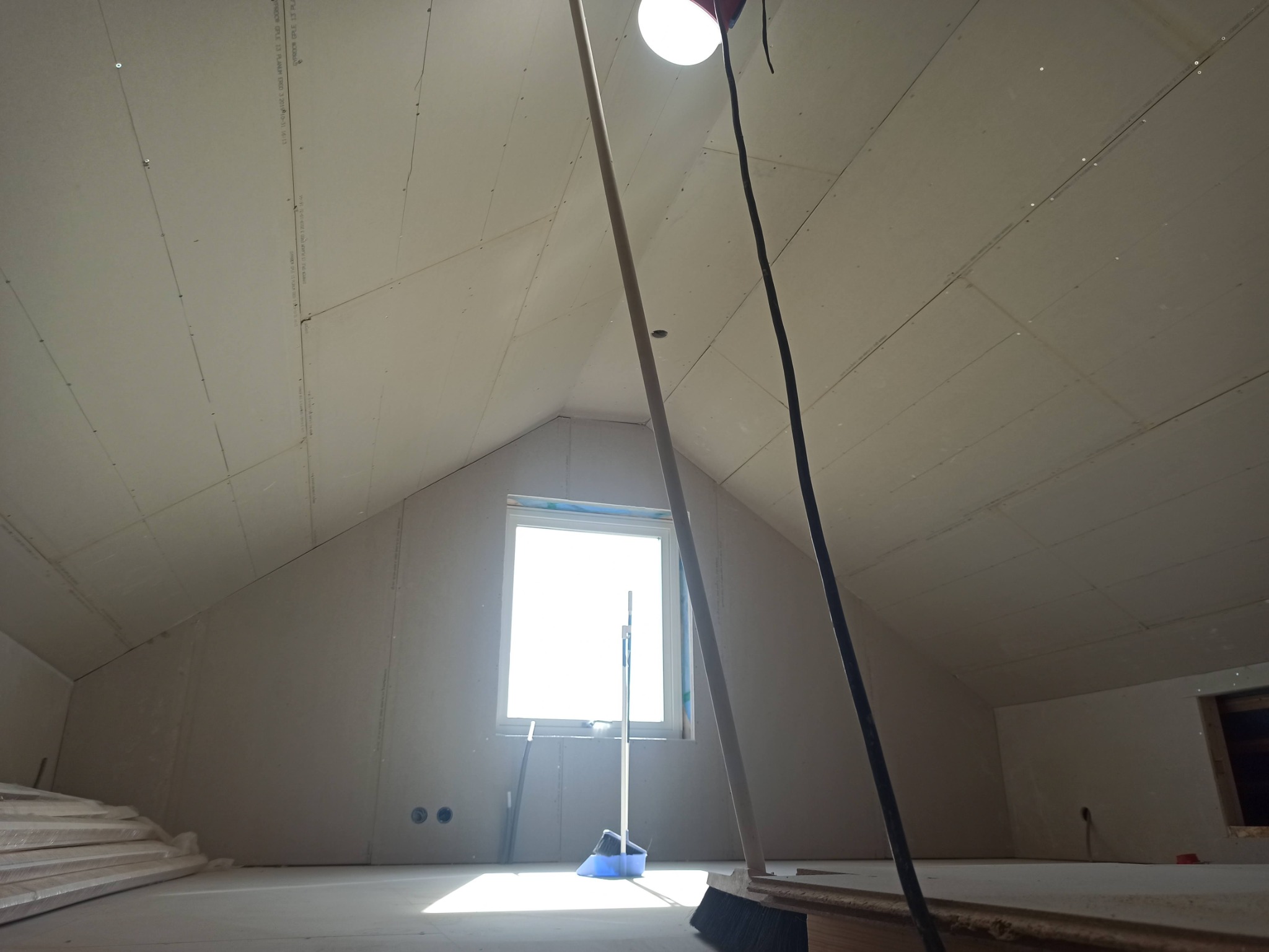 Poddasze w trakcie prac wykończeniowych, widoczne płyty gipsowo-kartonowe na ścianach i suficie, narzędzia i materiały budowlane, okno w szczycie.