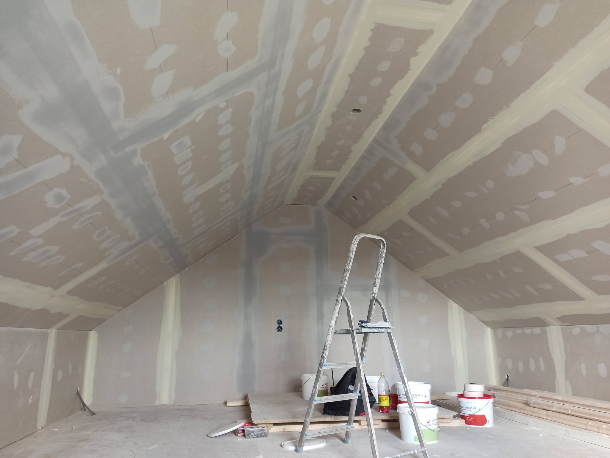 Poddasze w trakcie prac wykończeniowych, widoczne płyty gipsowo-kartonowe na ścianach i suficie, zaszpachlowane łączenia, aluminiowa drabina przystawna, wiadra z masą szpachlową i listwy...