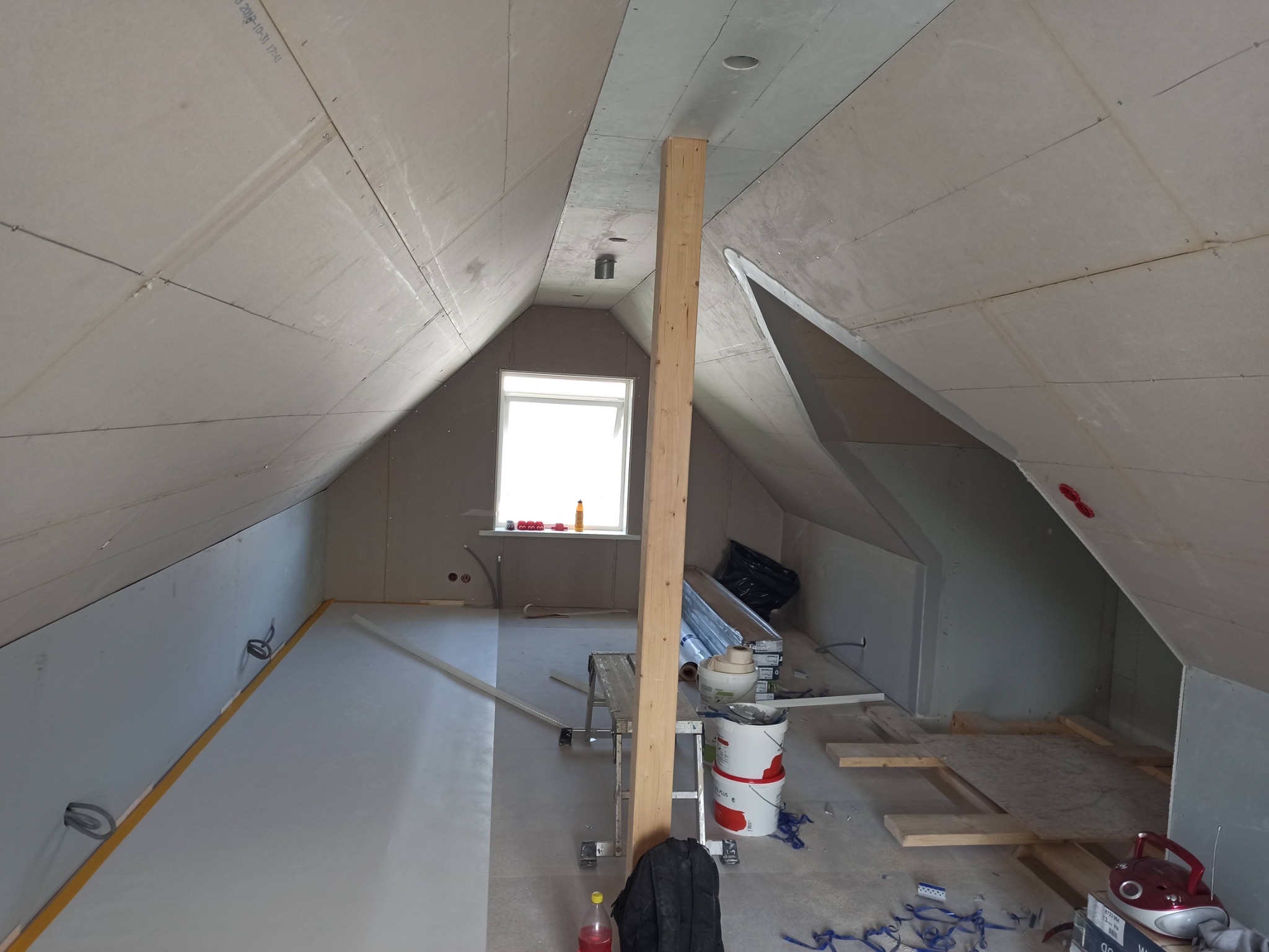 Poddasze w trakcie adaptacji: ściany obłożone płytami gipsowo-kartonowymi, zabezpieczona podłoga, widoczny słup drewniany, drabina, farby i narzędzia malarskie.