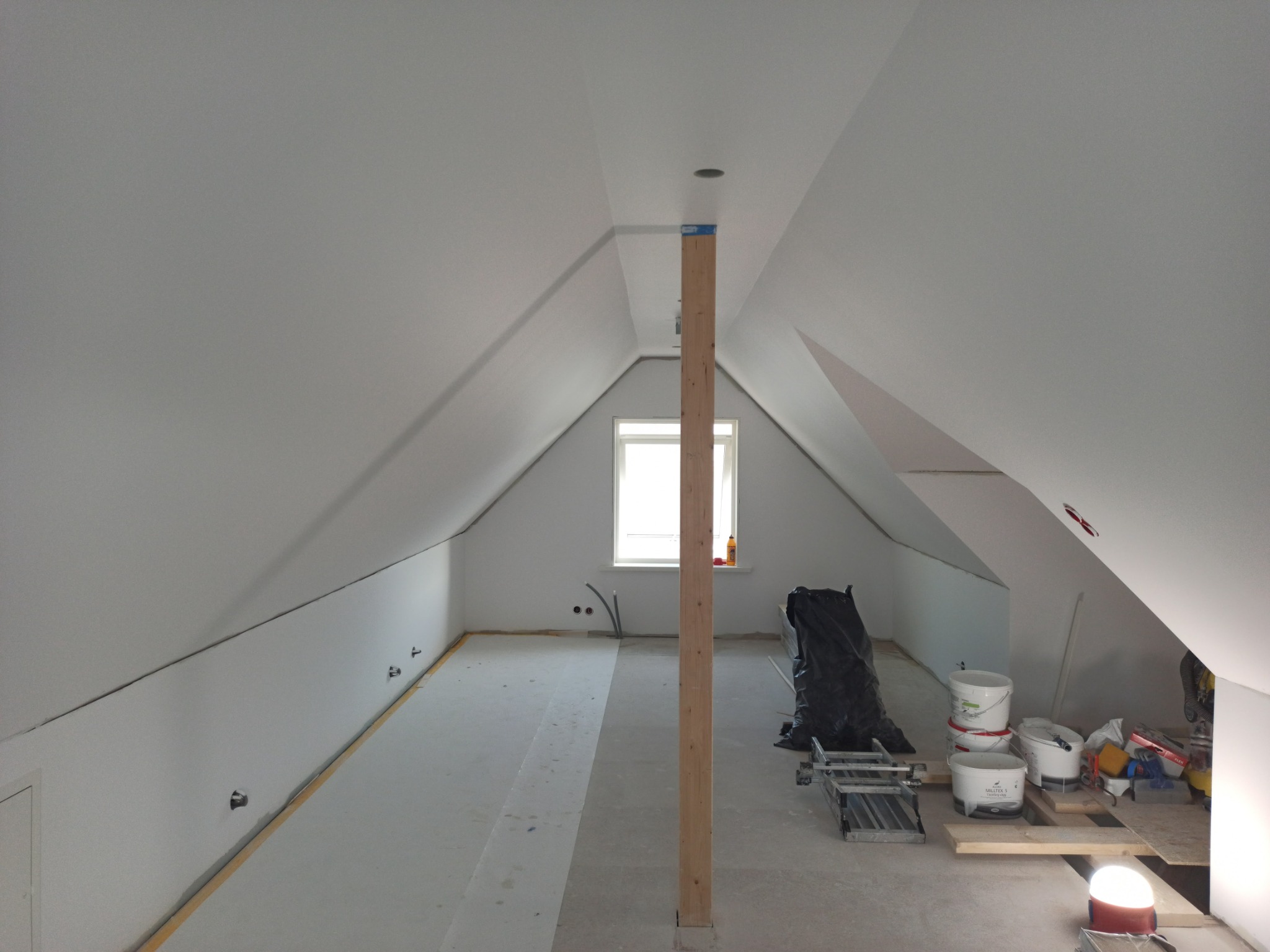 Poddasze w trakcie remontu: szpachlowane ściany i sufit, drewniany słup podtrzymujący, okno z widoczną butelką płynu, narzędzia i farby w tle.