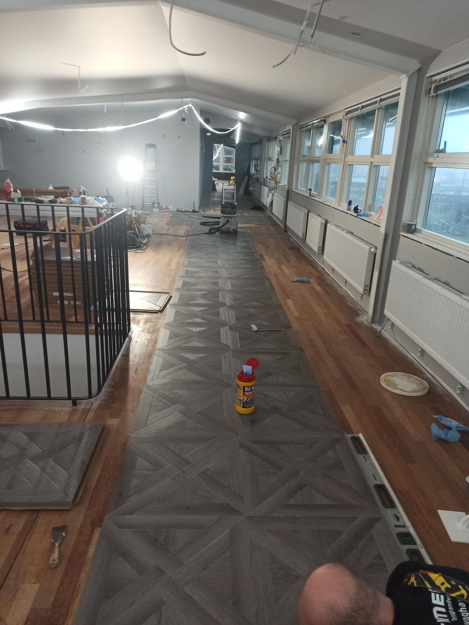 Układanie parkietu w geometryczny wzór w przestronnym wnętrzu, widoczne narzędzia i materiały budowlane, w tle okna i grzejniki.