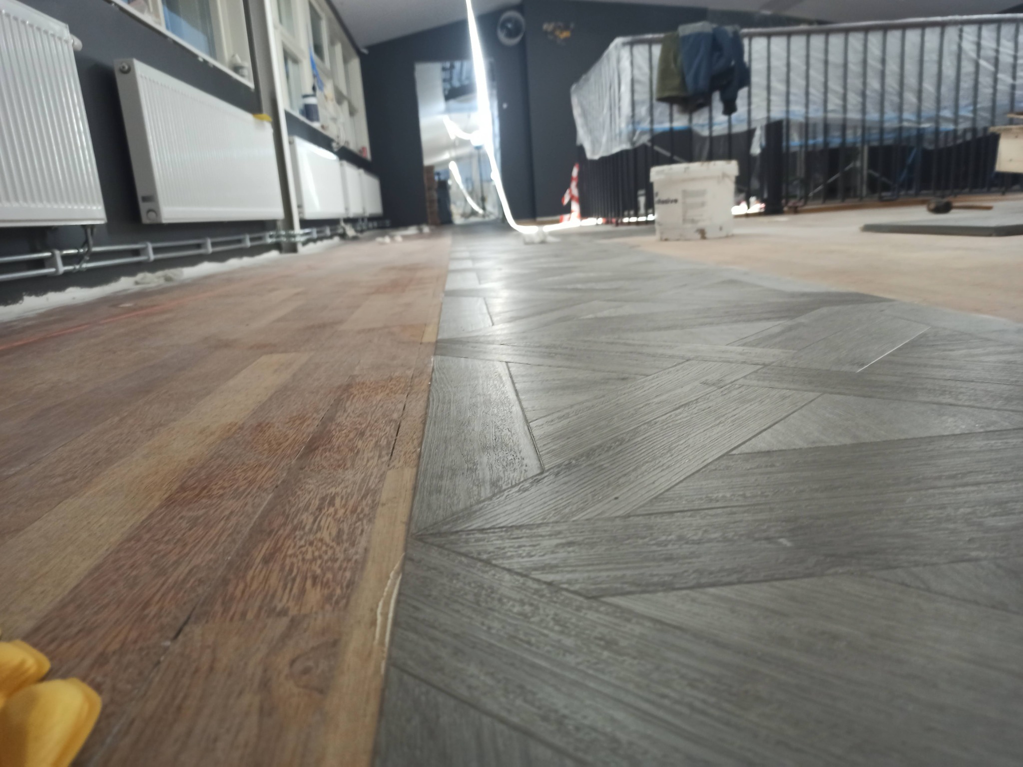 Podłoga w trakcie układania: parkiet w jodełkę w odcieniach szarości sąsiaduje z fragmentem surowej, drewnianej podłogi, widoczne grzejniki, wiadro farby i zabezpieczona folią balustrada w tle.