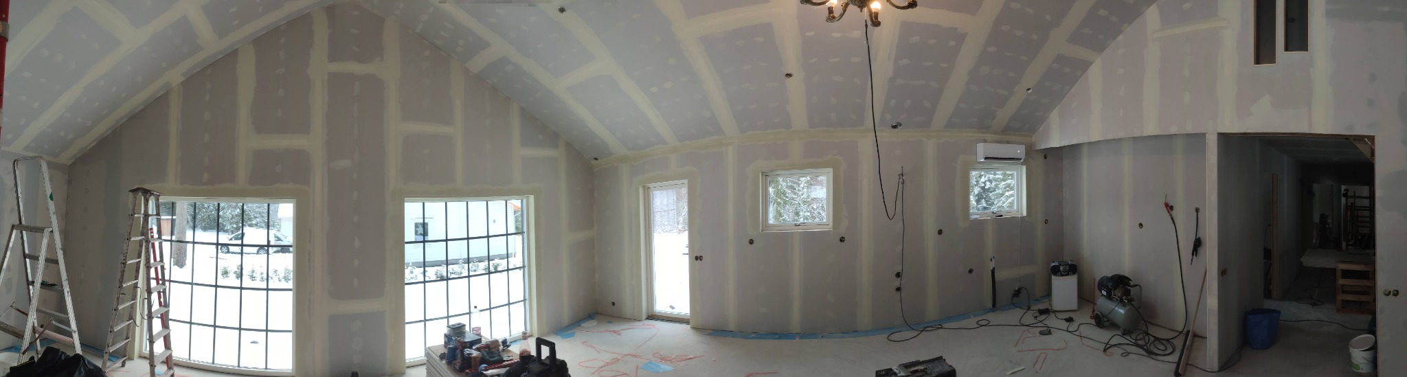 Szeroki kadr na wnętrze w trakcie szpachlowania; widoczne płyty gipsowo-kartonowe z nałożoną warstwą szpachli, okna z widokiem na zimowy krajobraz, oraz narzędzia i drabiny.