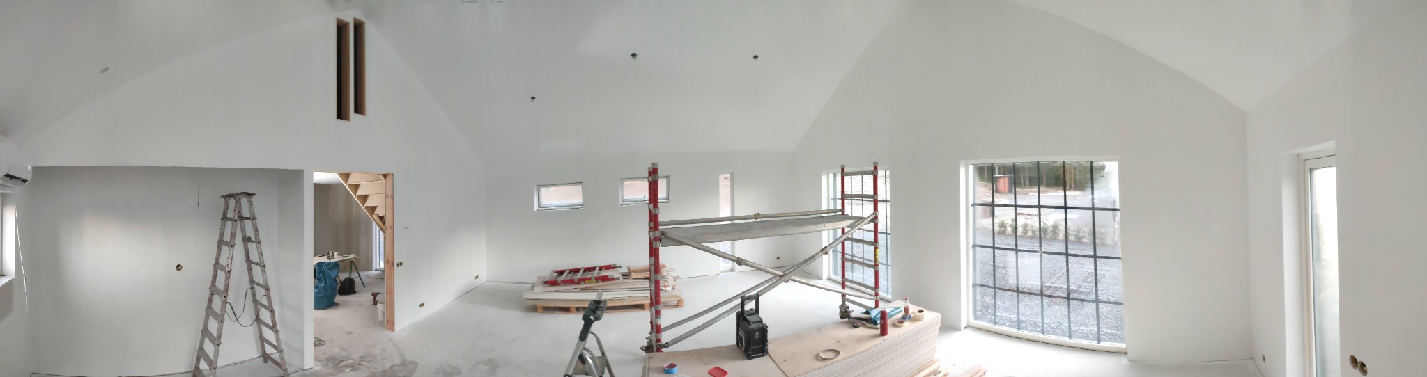 Wnętrze w trakcie prac wykończeniowych: ściany pomalowane na biało, widoczne rusztowanie, drabina, płyty gipsowe i narzędzia; surowe schody w tle.