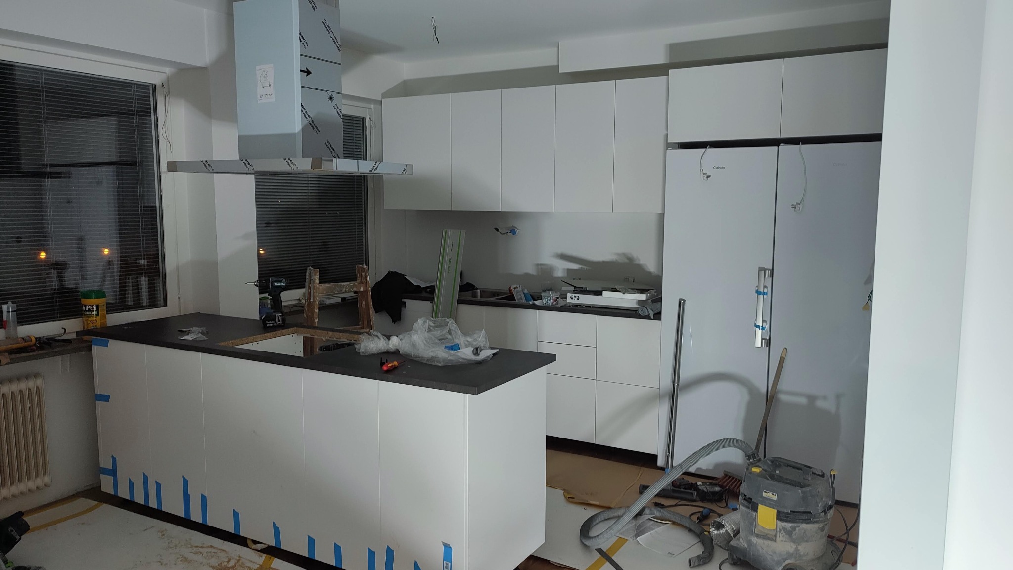 Wnętrze kuchni w trakcie montażu: białe szafki, wyspa z czarnym blatem zabezpieczona taśmą, sprzęt AGD, widoczny odkurzacz przemysłowy i drabina.