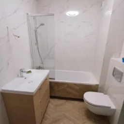 Nowoczesna łazienka po remoncie: białe marmurowe płytki, drewnopodobna podłoga w jodełkę, minimalistyczna umywalka i toaleta podwieszana.