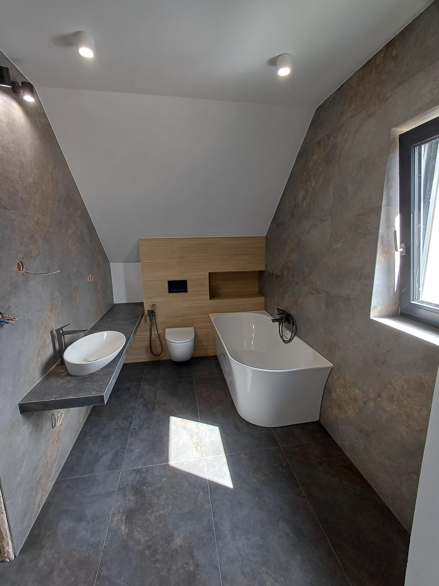 Nowoczesna łazienka na poddaszu z wanną wolnostojącą, umywalką na blacie z szarego kamienia i płytkami imitującymi kamień. Uwagę zwraca drewniana zabudowa toalety.