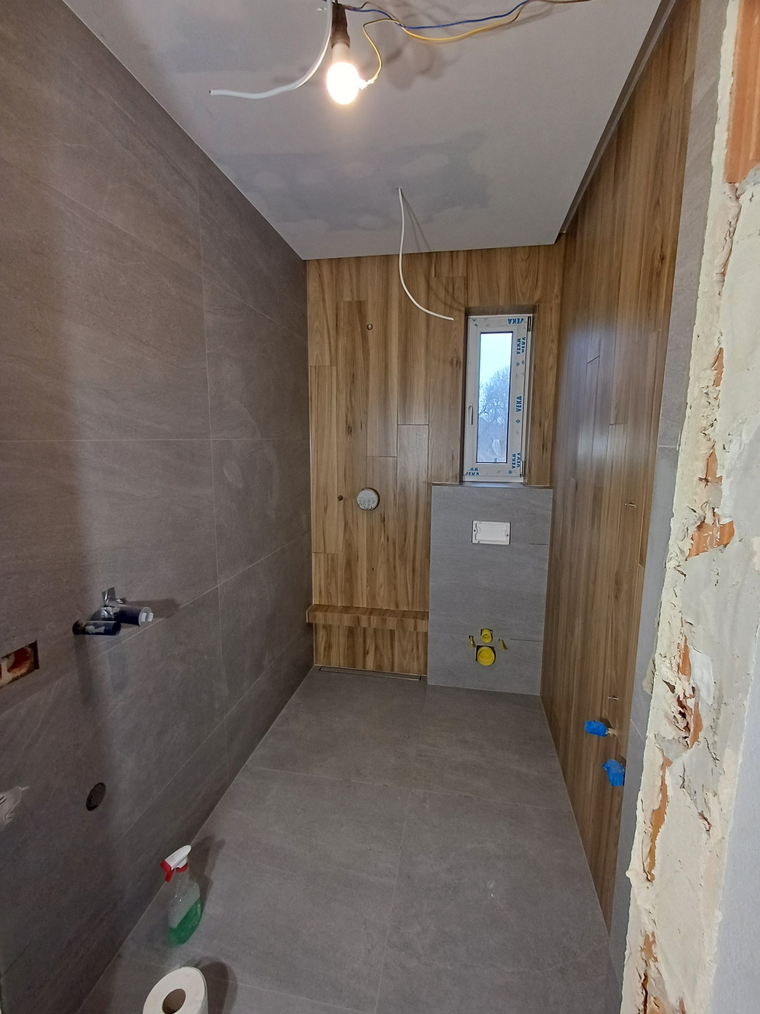 Remont łazienki: szare płytki na ścianach i podłodze, drewnopodobne panele na ścianach, okno w zabudowie, widoczne rury i instalacje, butelka z płynem na podłodze.