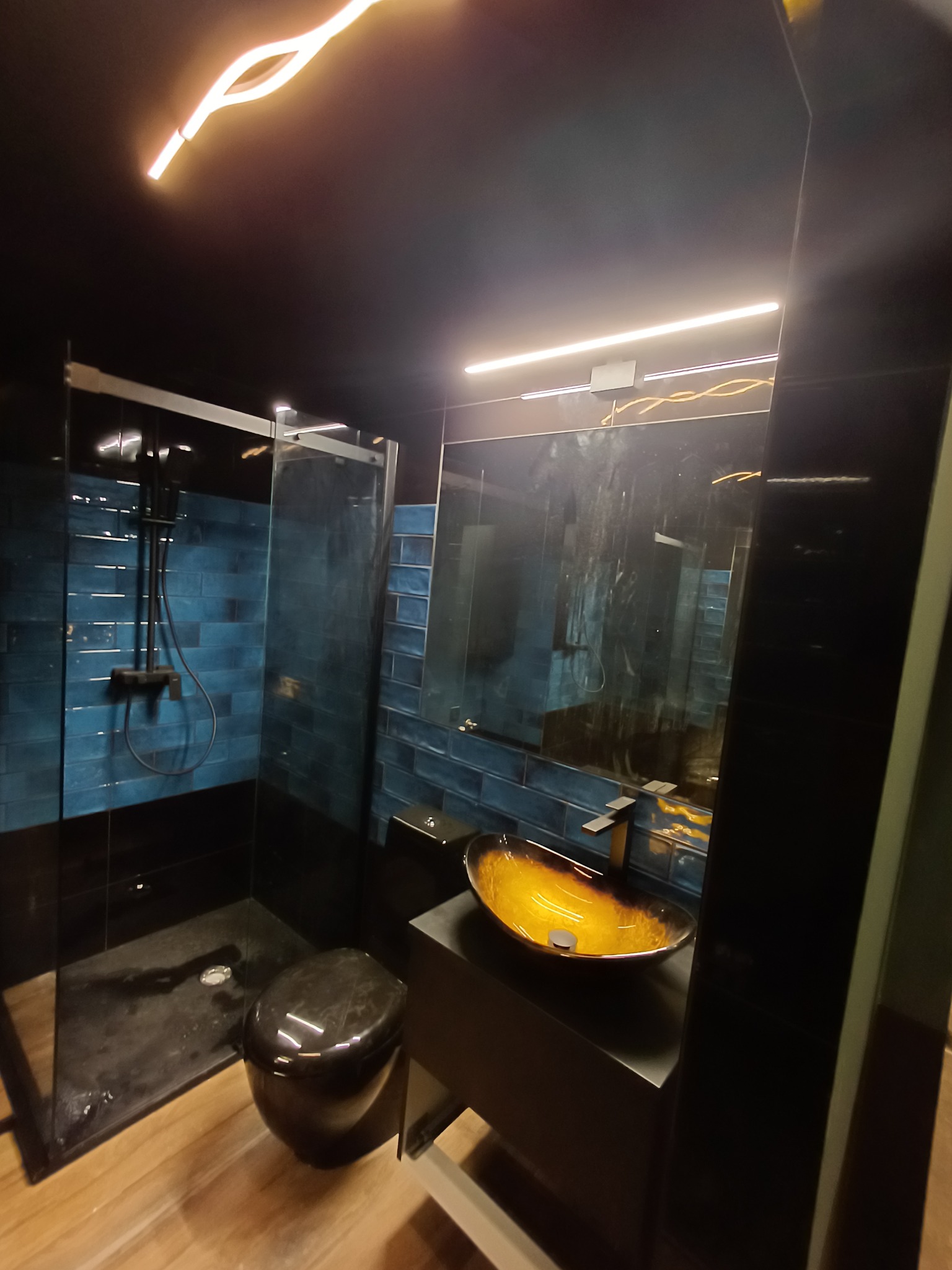 Nowoczesna łazienka z kabiną prysznicową typu walk-in, umywalką w odcieniach złota i czarną armaturą. Błękitne płytki na ścianie dodają wnętrzu koloru.