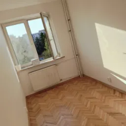 Pusty pokój z parkietem w jodełkę, świeżo po remoncie, z oknem wychodzącym na zieleń miejską i białymi ścianami, na parapecie stoi biały kubek i kartonowe pudełko.