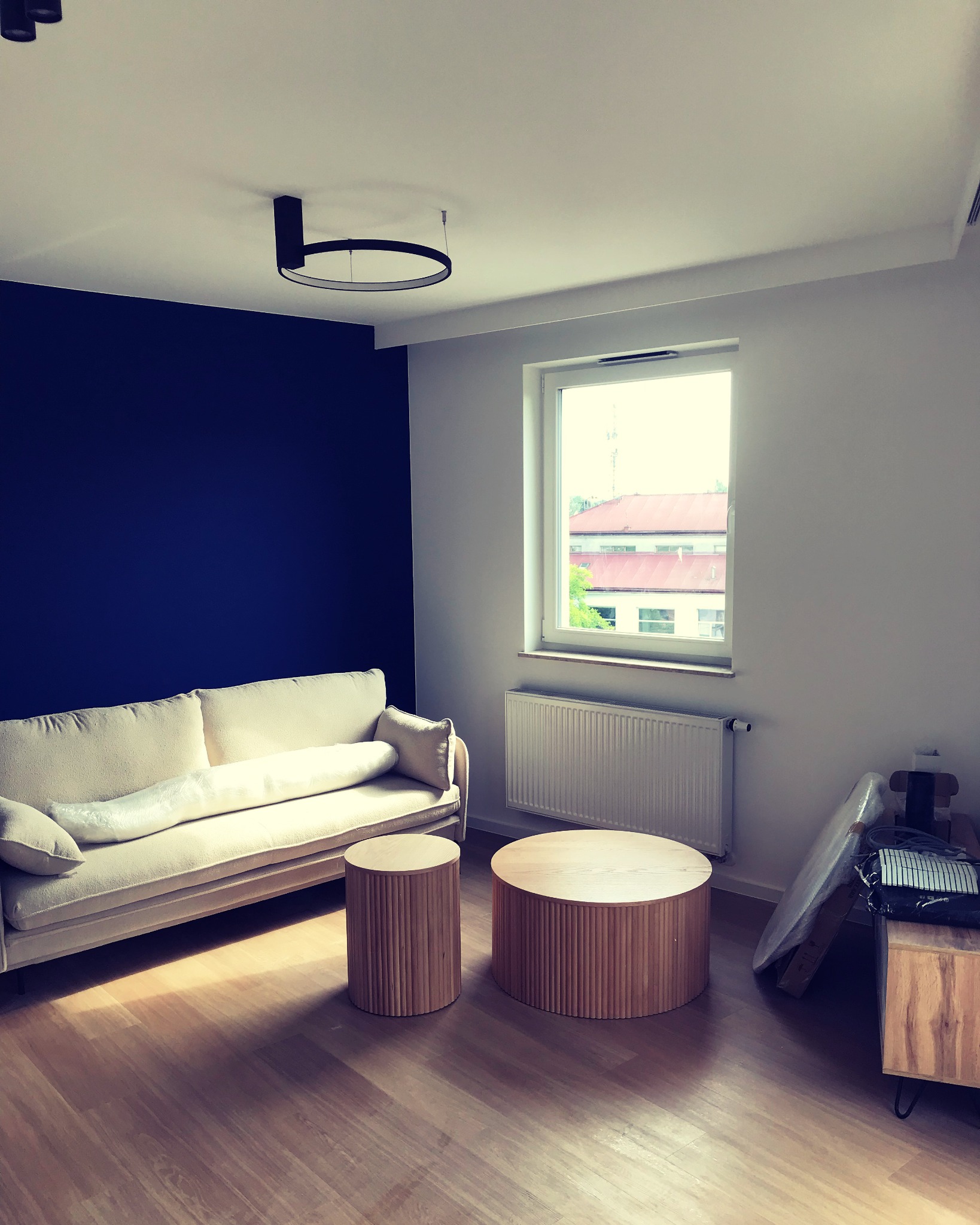 Salon po remoncie z granatową ścianą, jasną podłogą, kremową sofą i drewnianymi stolikami, widoczne akcesoria w folii obok grzejnika.