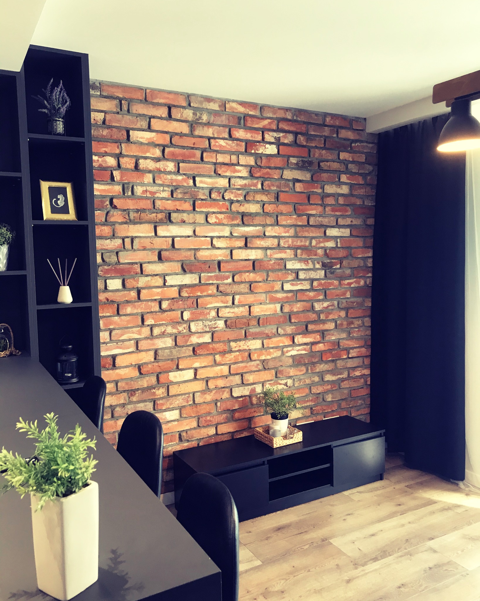 Wnętrze pokoju z dekoracyjną ścianą z czerwonej cegły, czarnymi meblami i jasną drewnianą podłogą, oświetlone lampą wiszącą.