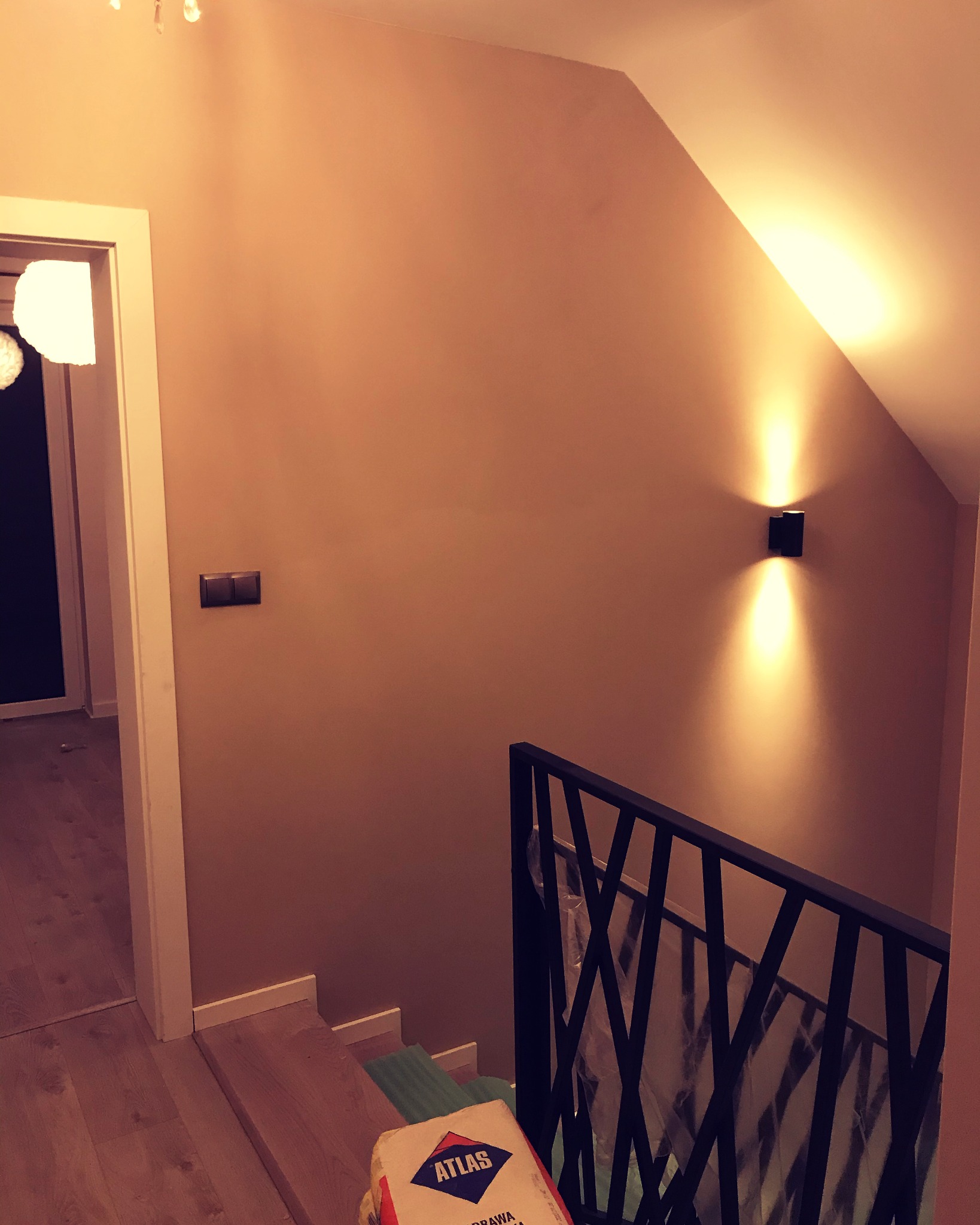 Świeżo pomalowana ściana w odcieniach beżu z nowoczesnym oświetleniem ściennym, widoczny fragment schodów z workiem zaprawy Atlas u podnóża, sugerujący prace wykończeniowe.